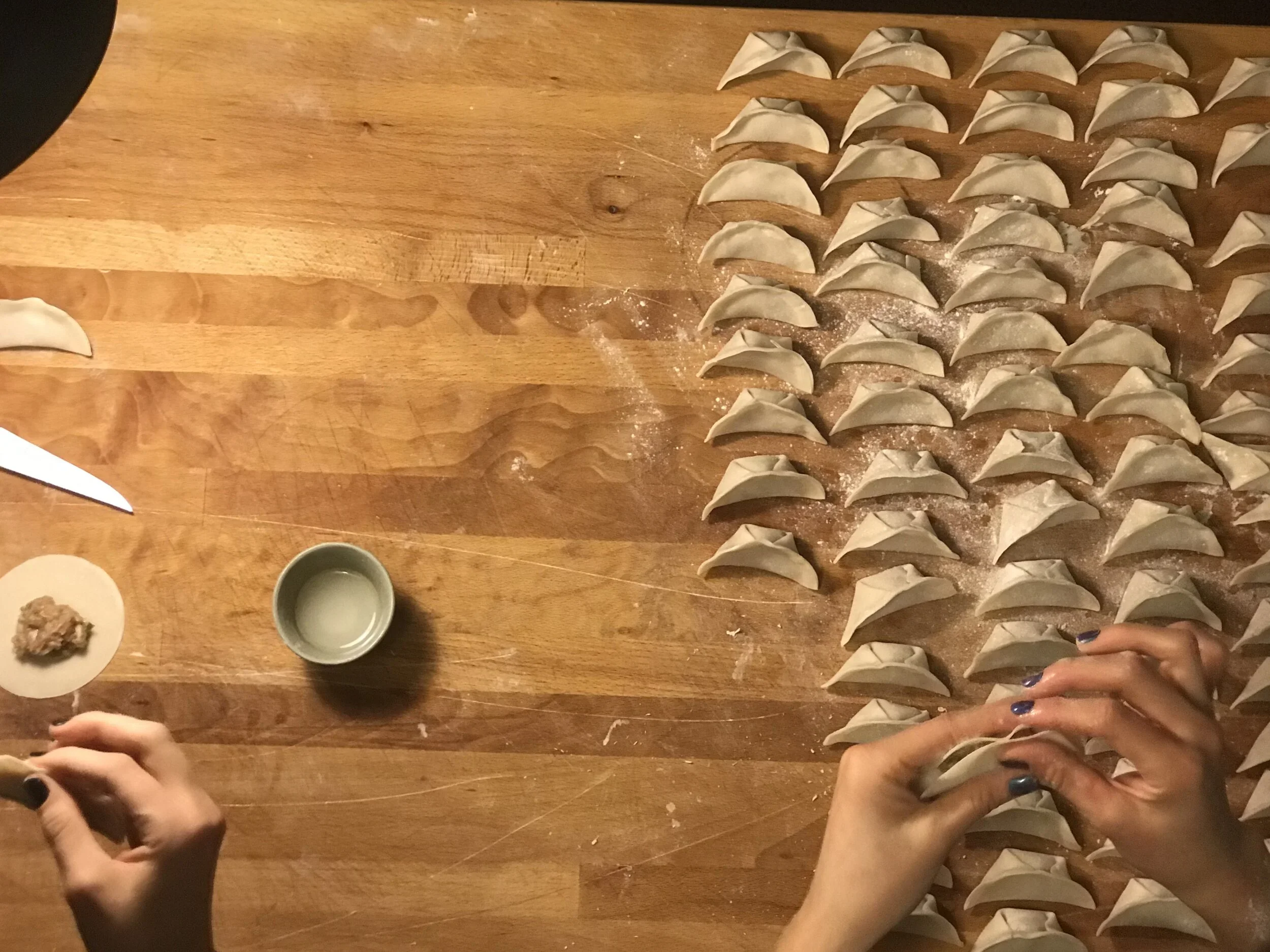 Hands folding homemade dumplings on a wooden table with a knife, a small bowl, and a plate of filling nearby.