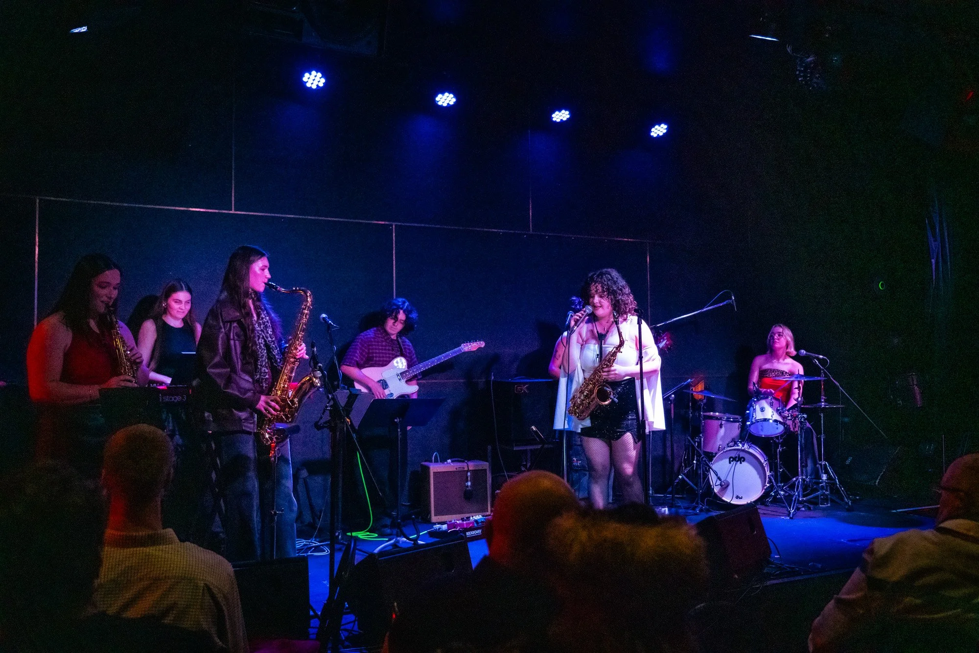A music concert with a band performing on stage. The band includes a female singer with curly hair, playing the saxophone, a drummer, a guitarist, and three female musicians playing saxophones and keyboard, illuminated by stage lights, with audience members watching.