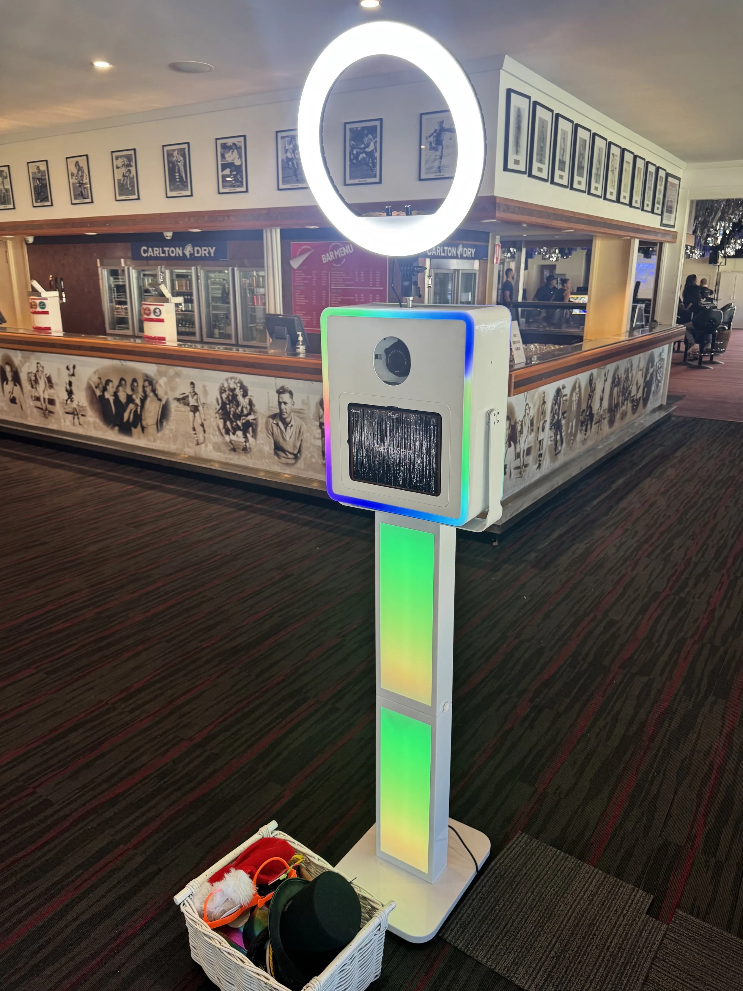 A photo booth with a ring light, facial recognition camera, and LED lights in a corridor of a restaurant or entertainment venue. There is a basket of hats and accessories at the base of the booth.