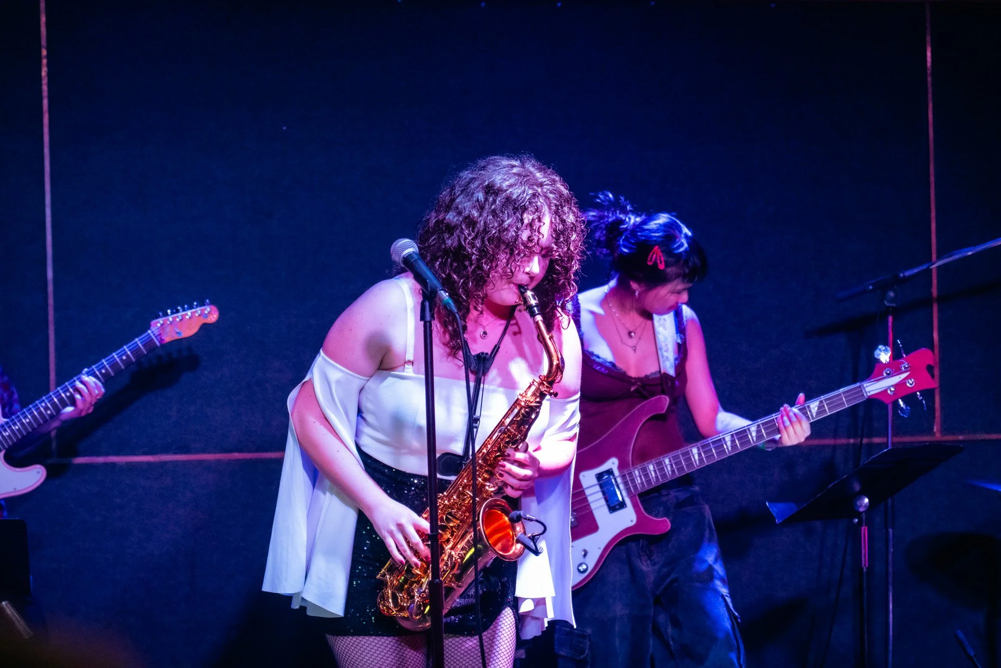 Two women playing musical instruments on stage, with one woman playing a saxophone in the spotlight and the other playing an electric guitar in the background.