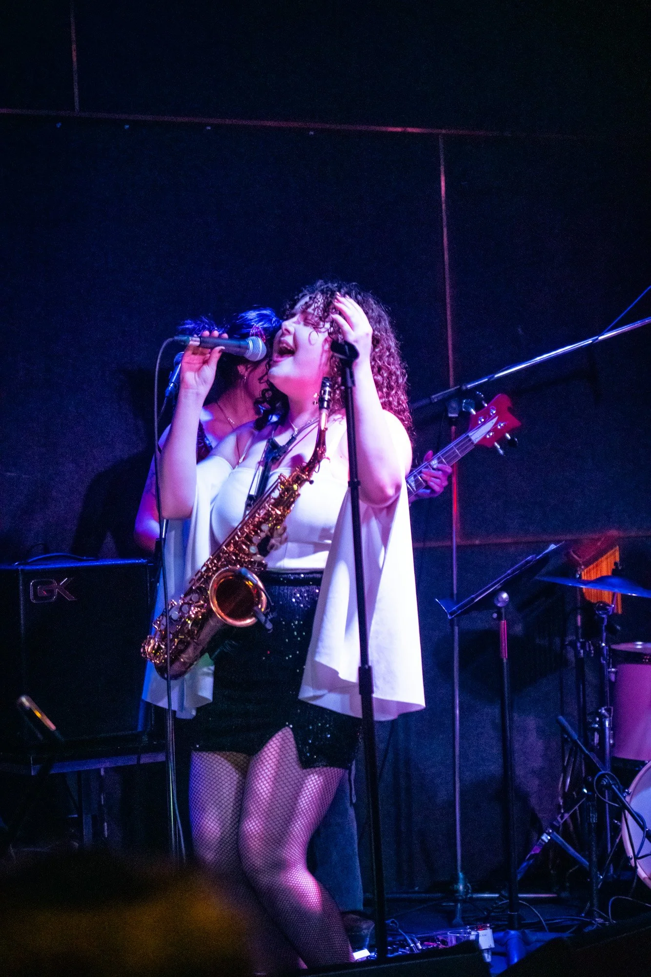 Andrea singing into a microphone with a saxophone hanging around her neck on stage during a live music performance, with a musician playing bass guitar in the background.