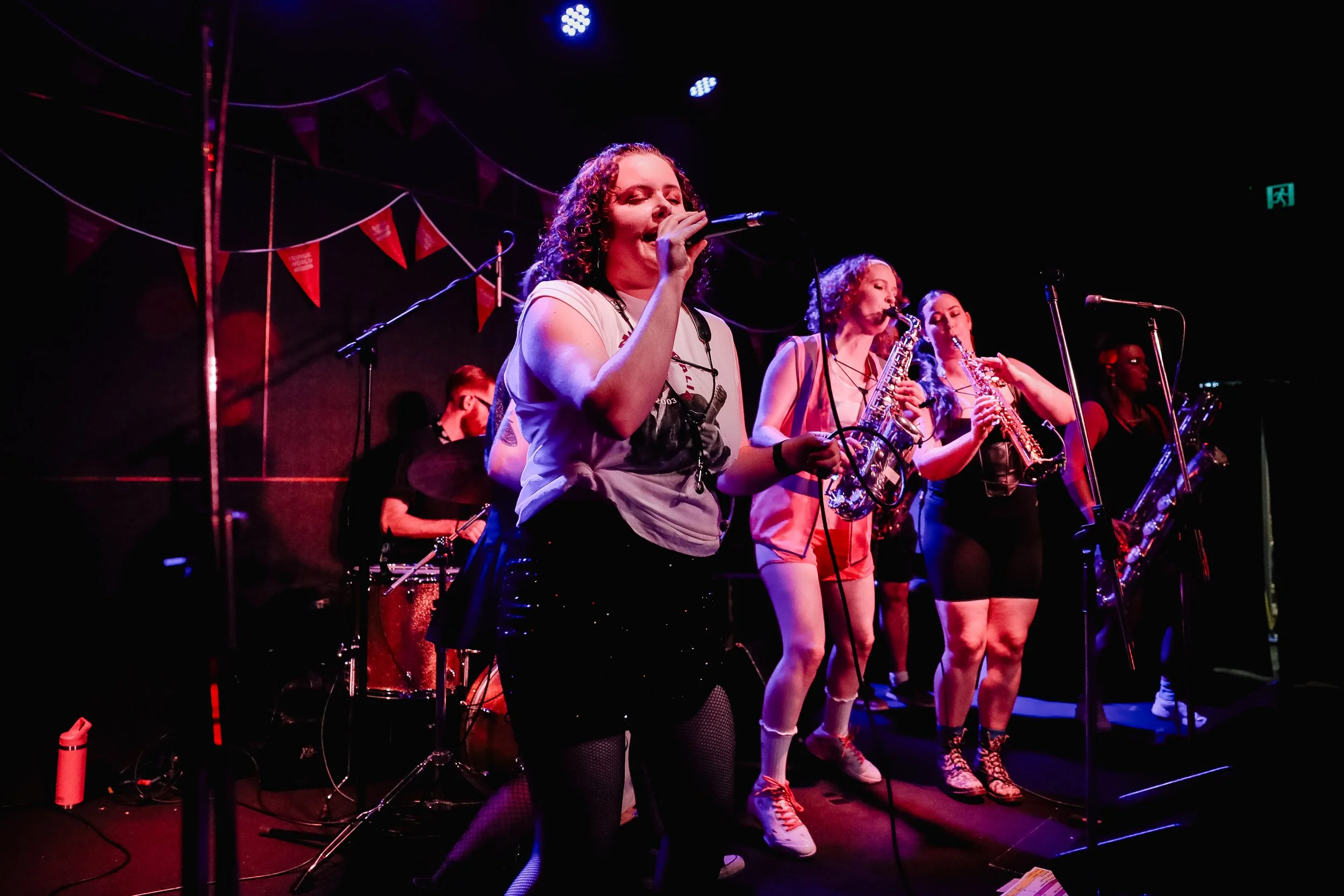 A musical group performs on stage, featuring a singer at the front with curly hair, wearing a casual white t-shirt, and four musicians behind playing saxophones, with colorful stage lighting and decorations.