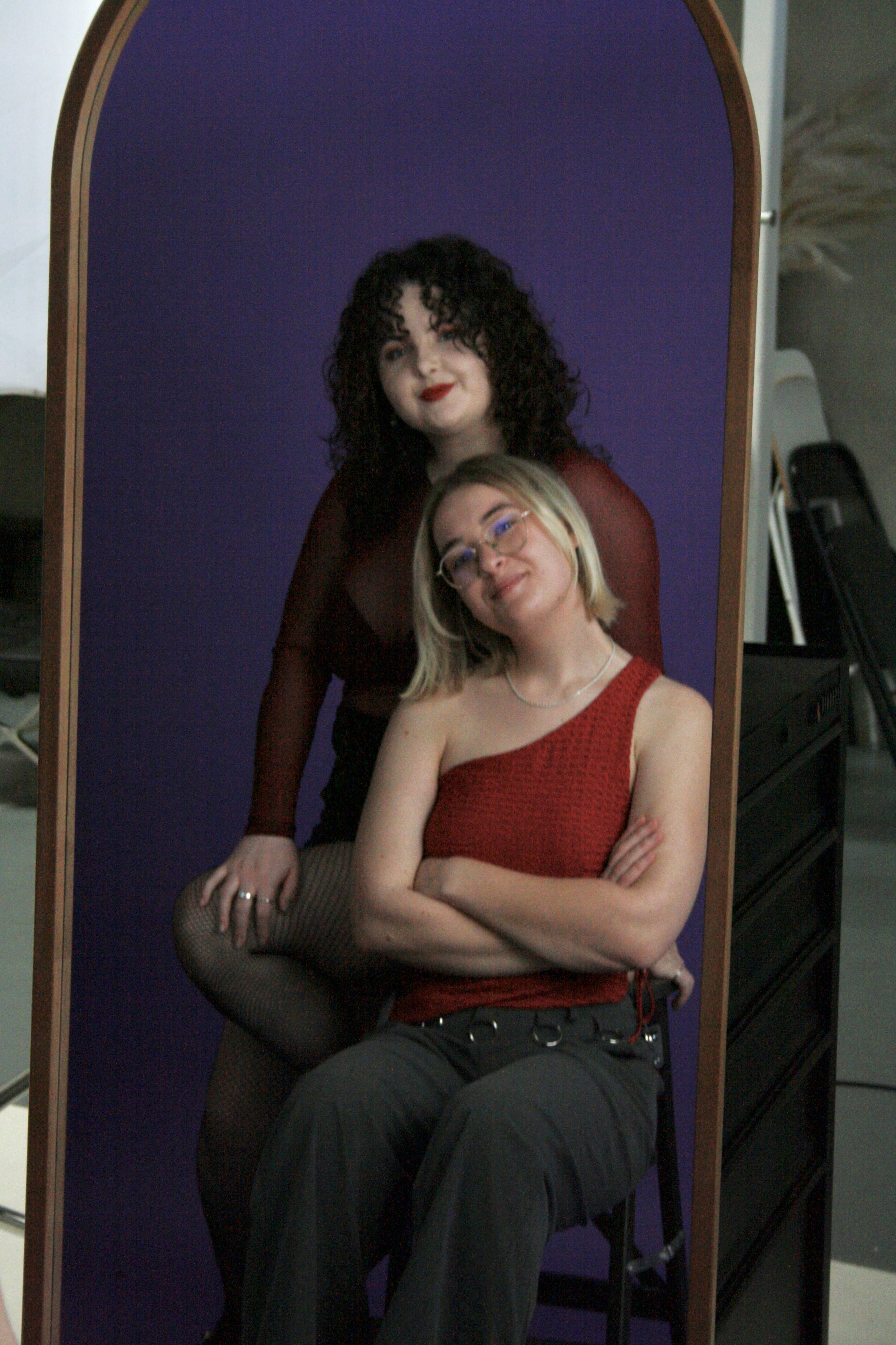 A mirror reflecting Andrea and her co-collaborator for Echo of Aphrodite posing together, Martha is sitting with arms crossed and the Andrea is standing behind with one leg raised, both smiling and dressed in stylish clothing.