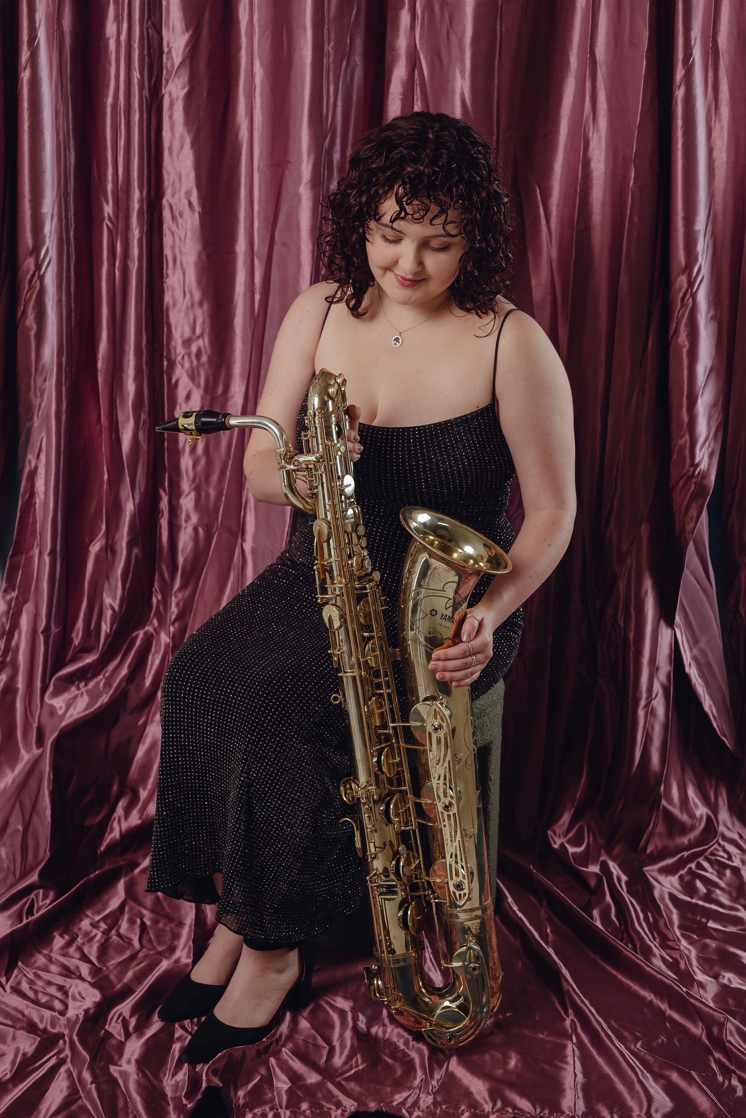 Andrea Jordan-Keane with her curly dark hair out and in a black dress holding a golden Baritone saxophone in front of a pink satin curtain.
