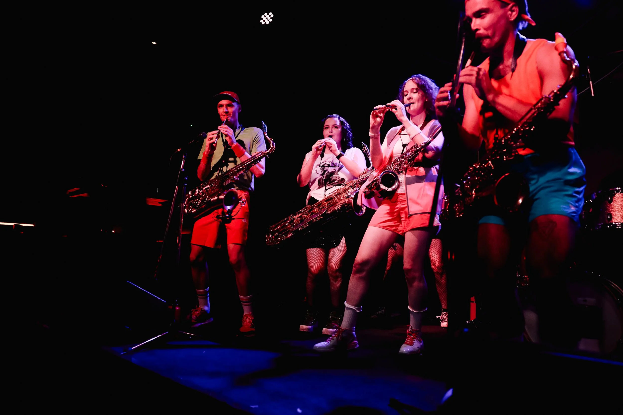 A band of four young musicians performing on stage, playing saxophones and a flute, dressed in colorful casual clothes with shorts, t-shirts, and sneakers, under stage lighting.