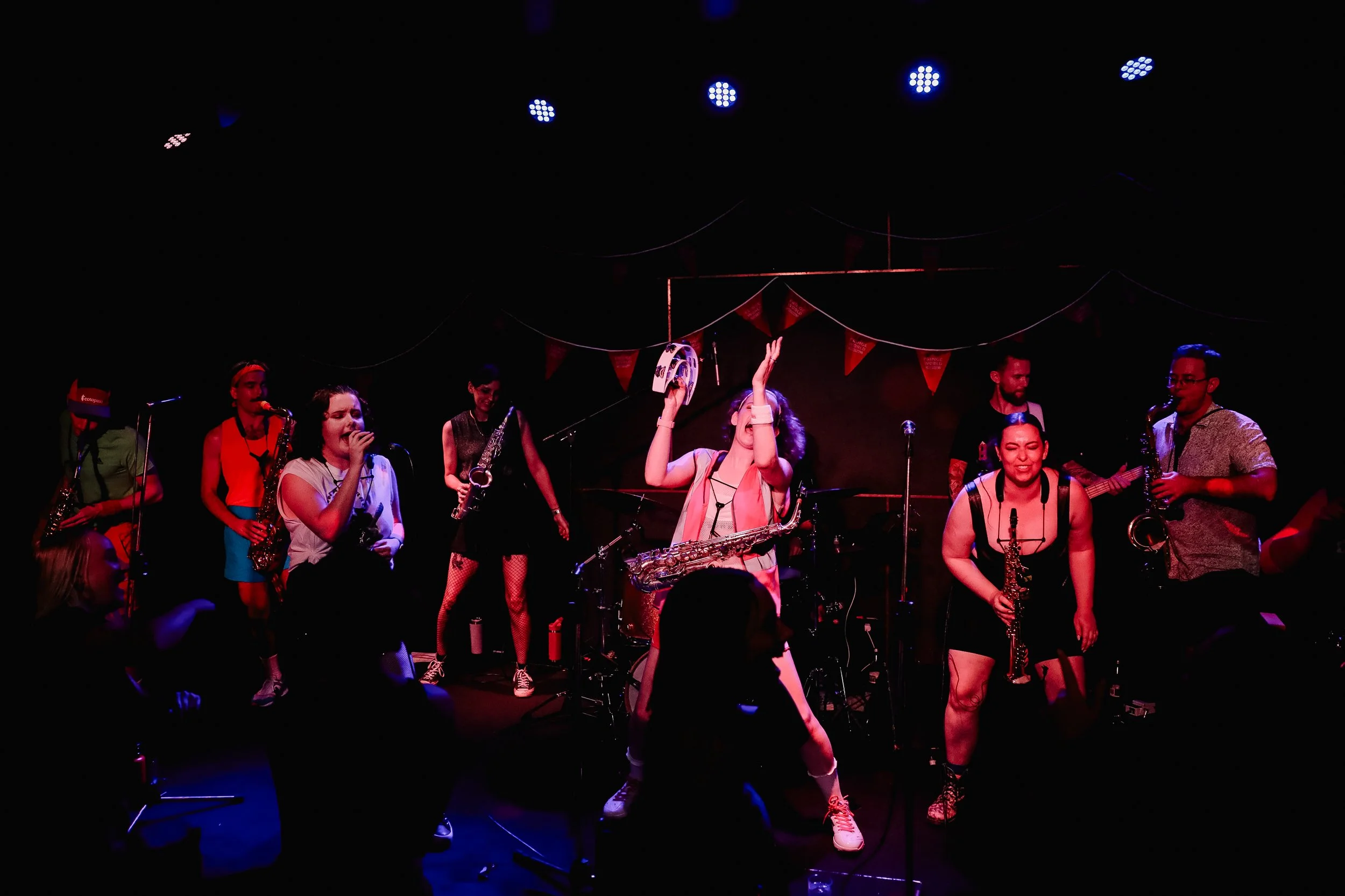 A live band performance on stage with several musicians, some singing and others playing instruments, in a dimly lit venue with colorful stage lights and red banners hanging in the background.