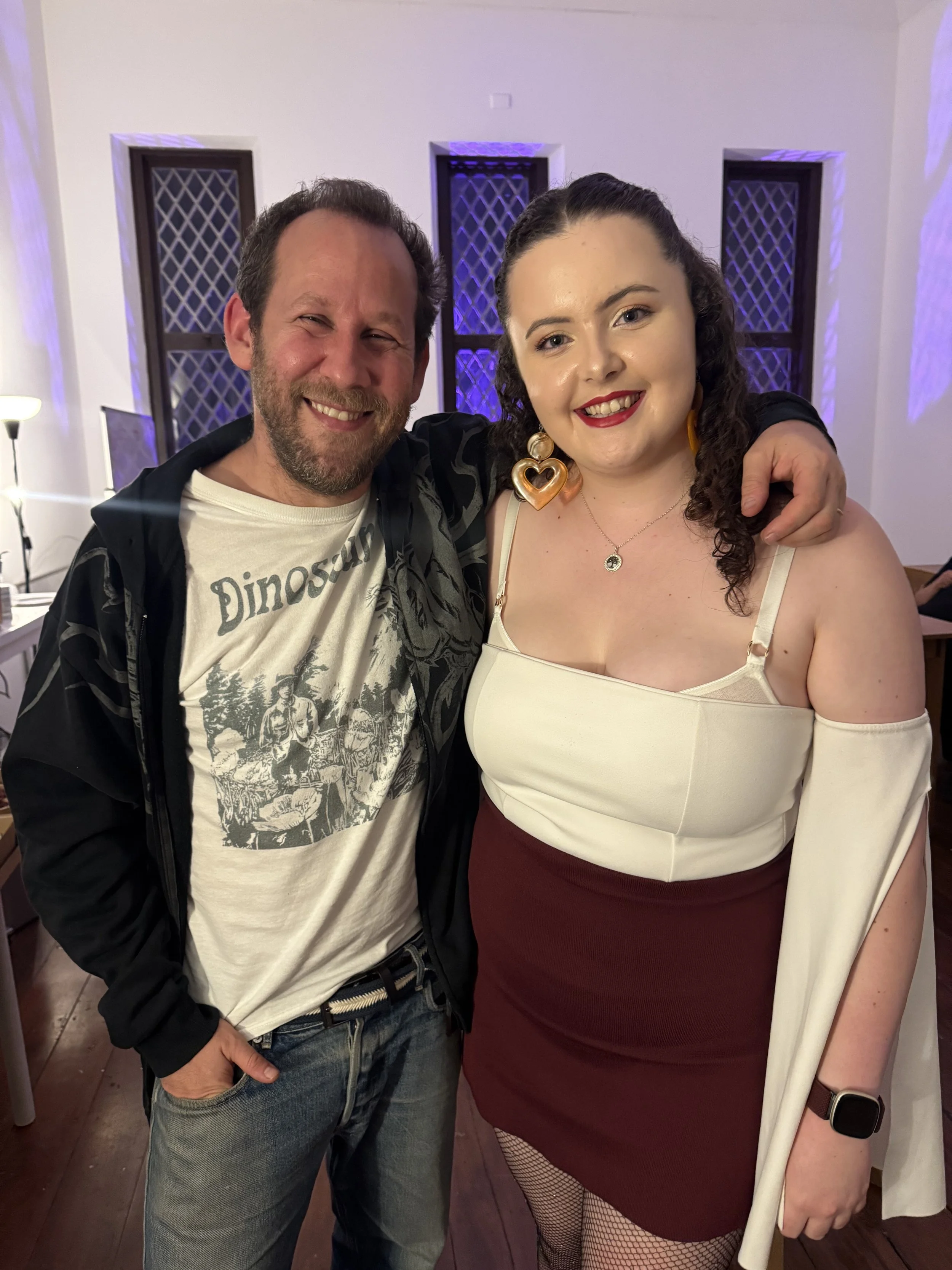 Ben Lee and Andrea Jordan-Keane smiling and posing together with arms around each other in a room with purple lighting and lattice windows.
