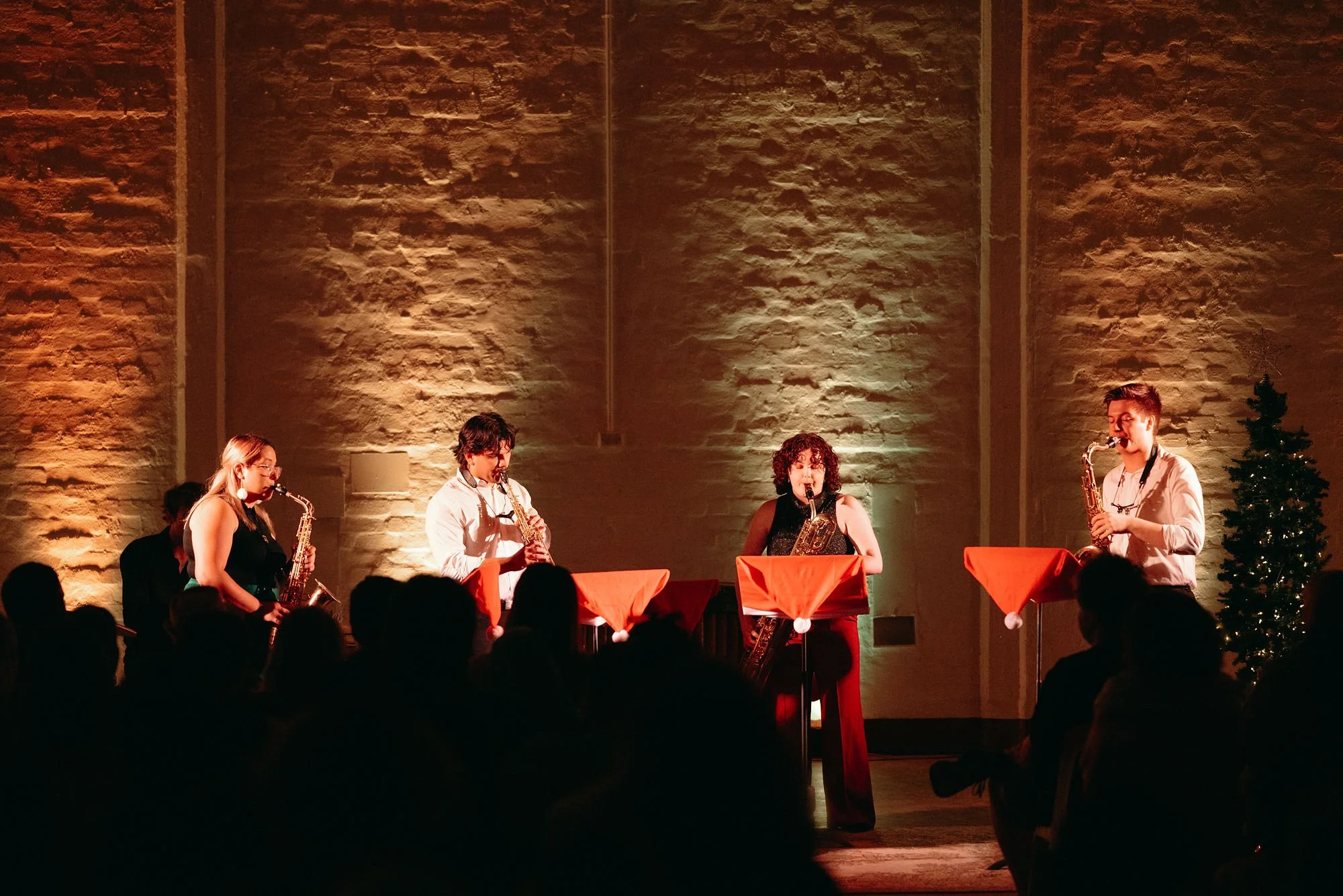 Five musicians playing saxophones on stage during a concert, with Christmas-themed decorations and a lit background.