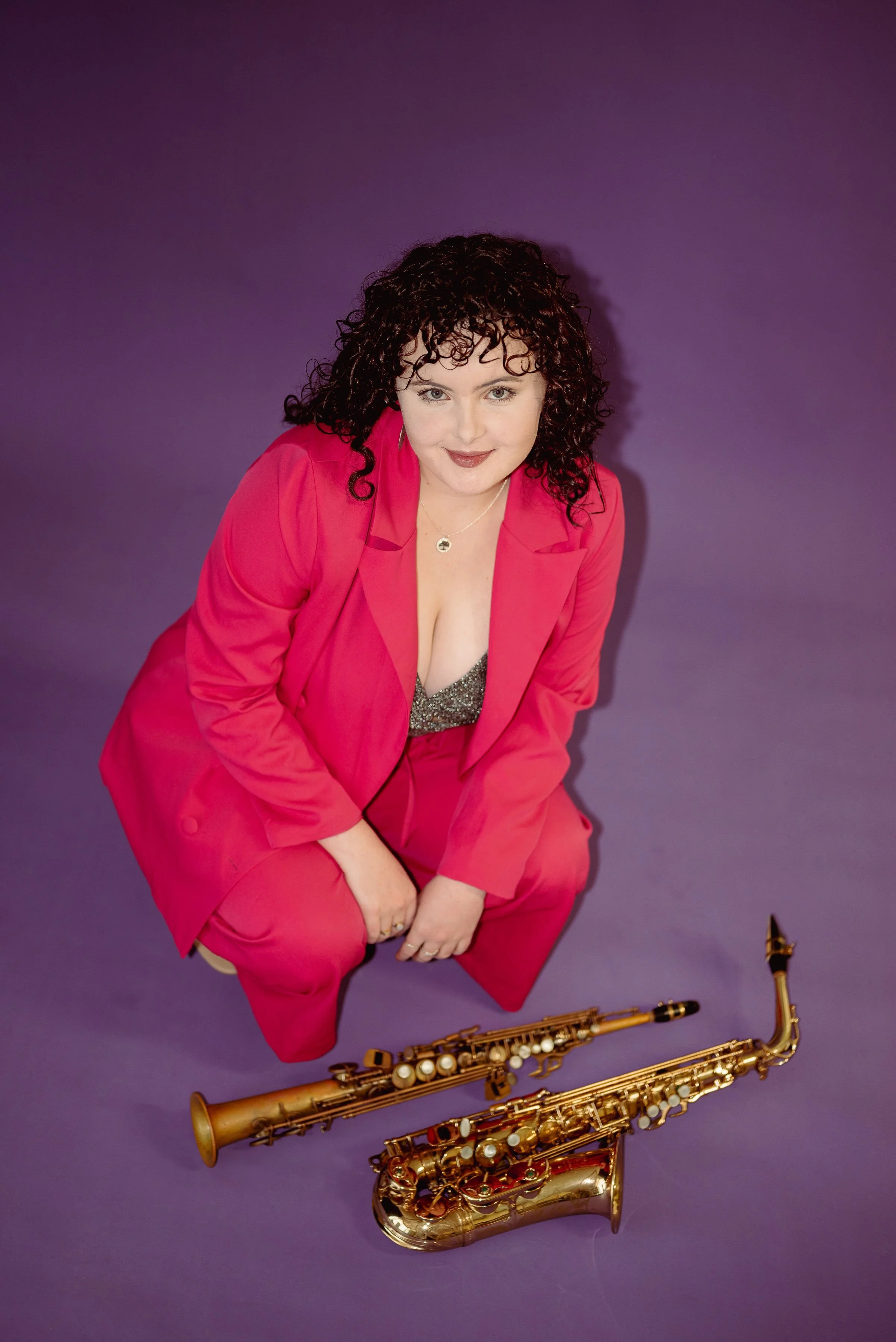 A woman with curly dark hair, wearing a bright pink suit, kneeling on a purple surface with two gold soprano and alto saxophones laying in front of her, against a matching purple background.