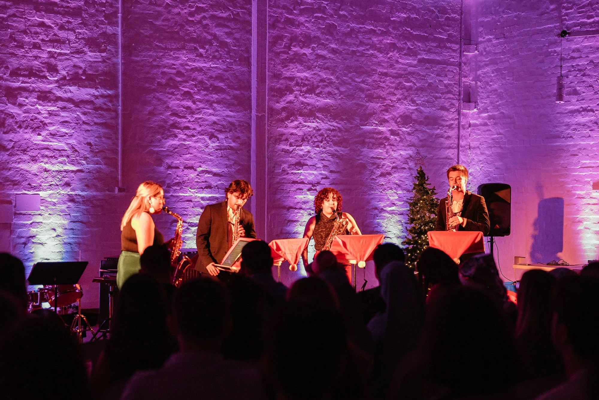 Musicians performing on stage with purple lighting and a small Christmas tree in background, audience watching.