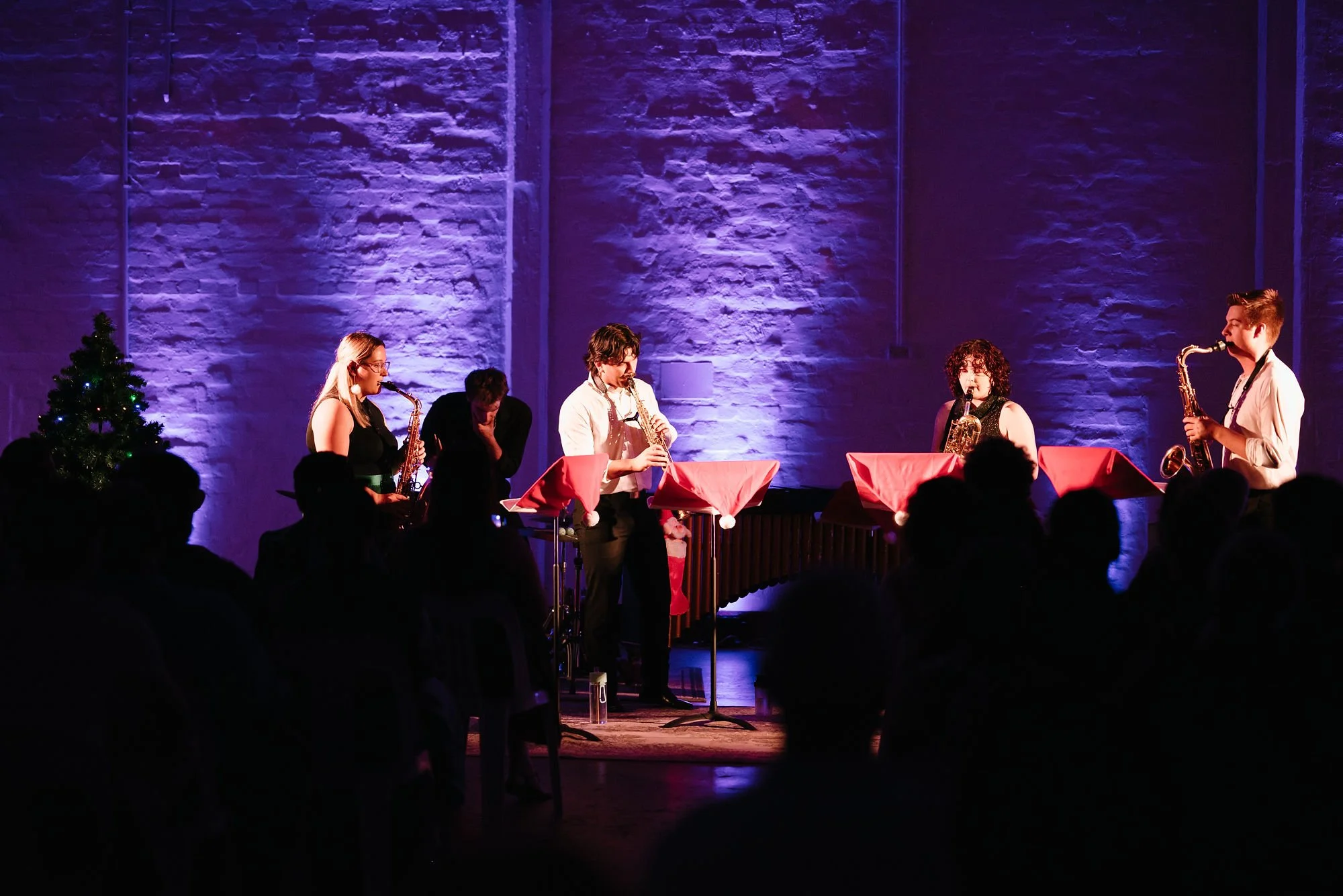 Symbios Saxophone Quartet performing on stage with purple lighting, in front of an audience, with a Christmas tree on the left.