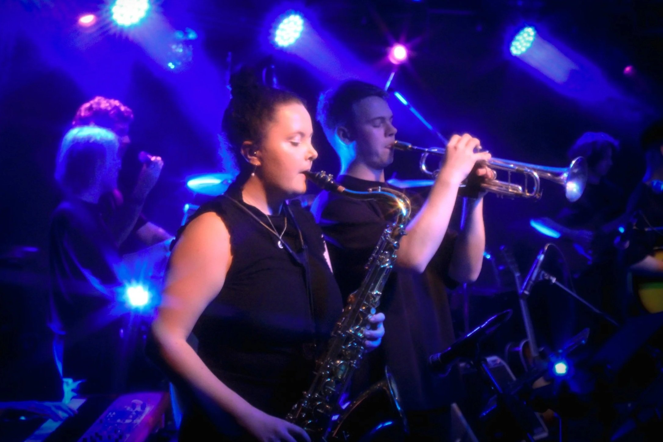 Musicians performing on stage with blue and purple lighting, including a woman playing the saxophone and a man playing the trumpet.