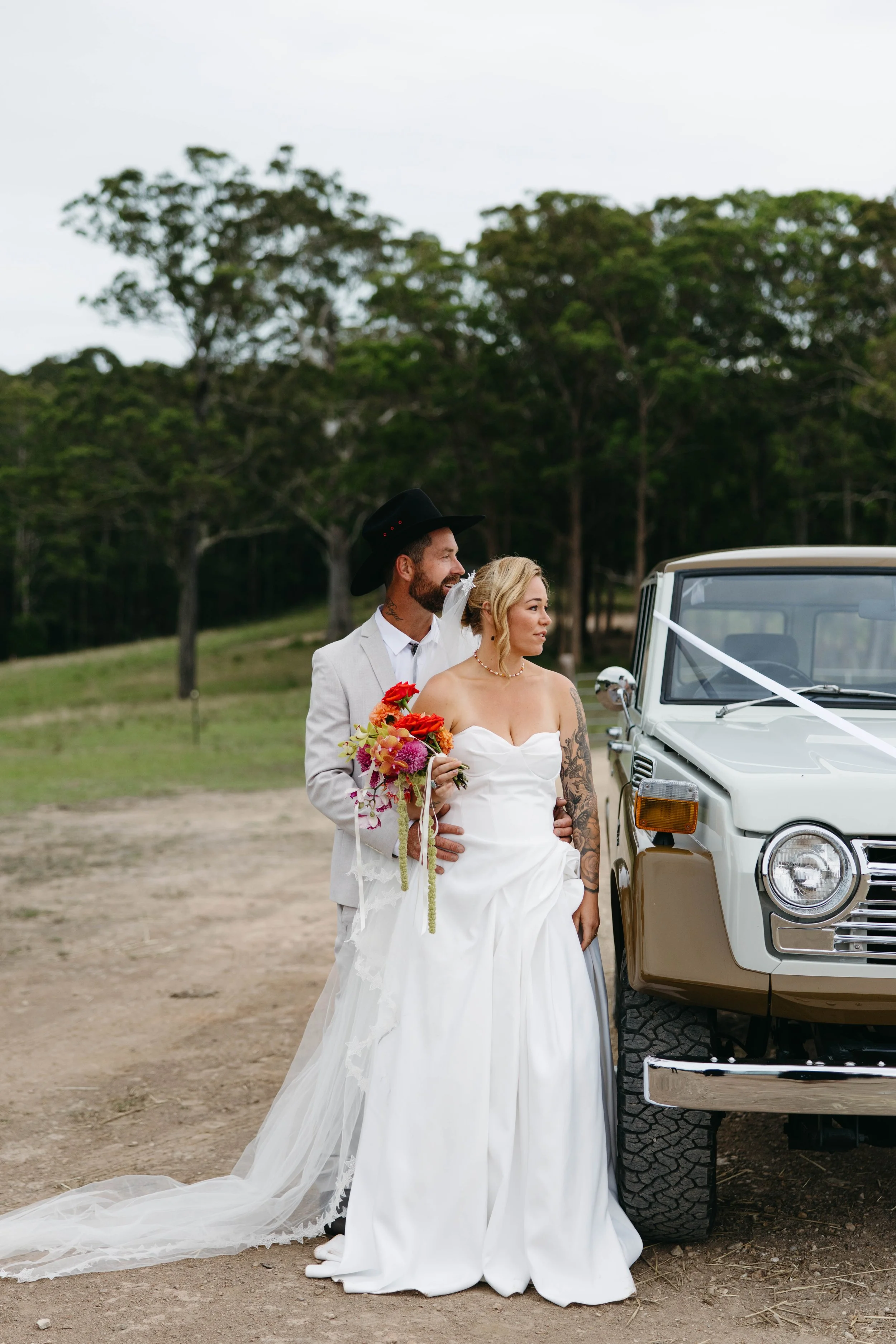 A bride and groom in wedding attire standing outdoors near a vintage vehicle, with trees and an overcast sky in the background.