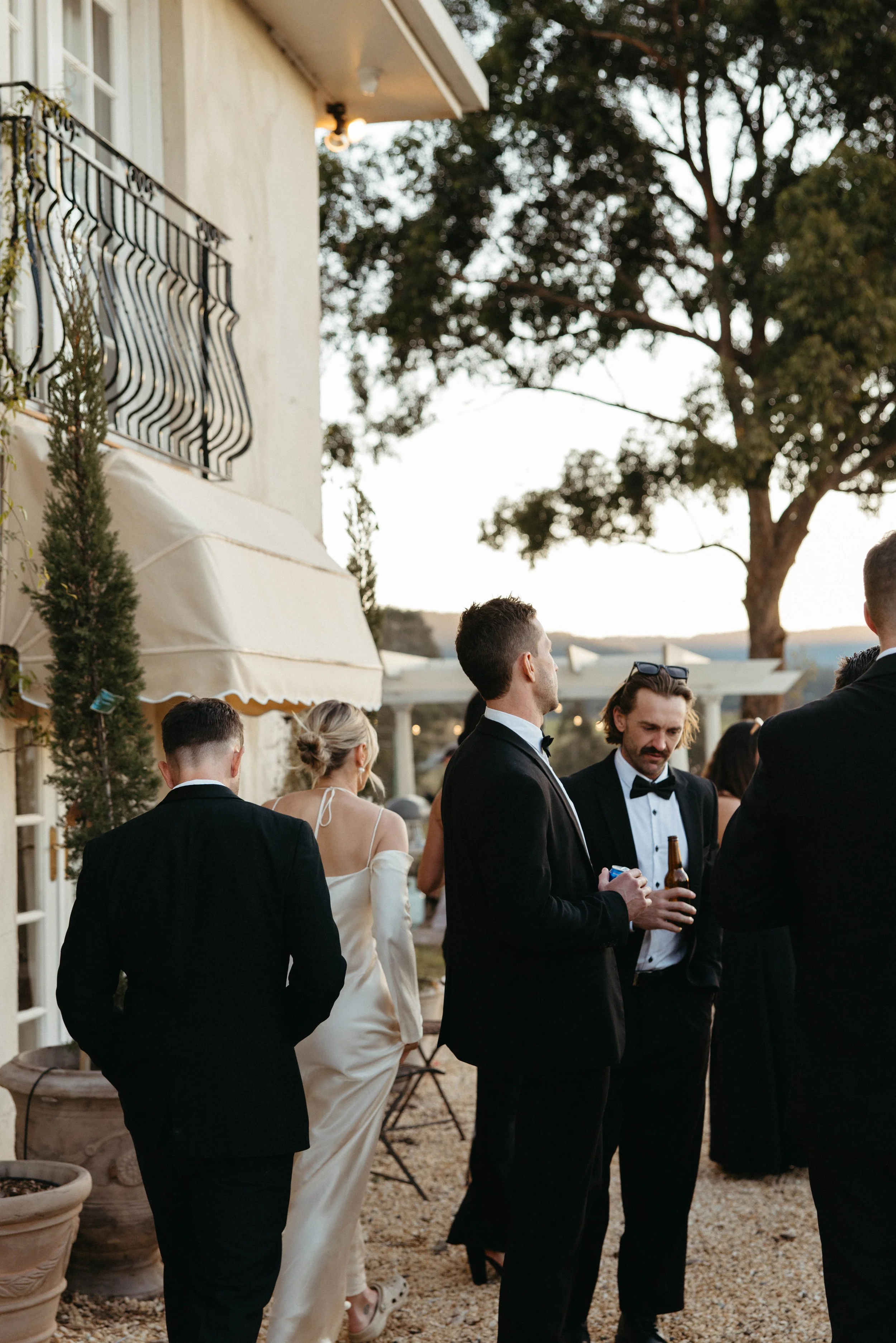 Wedding guests in black tie mingling outside a white villa at dusk