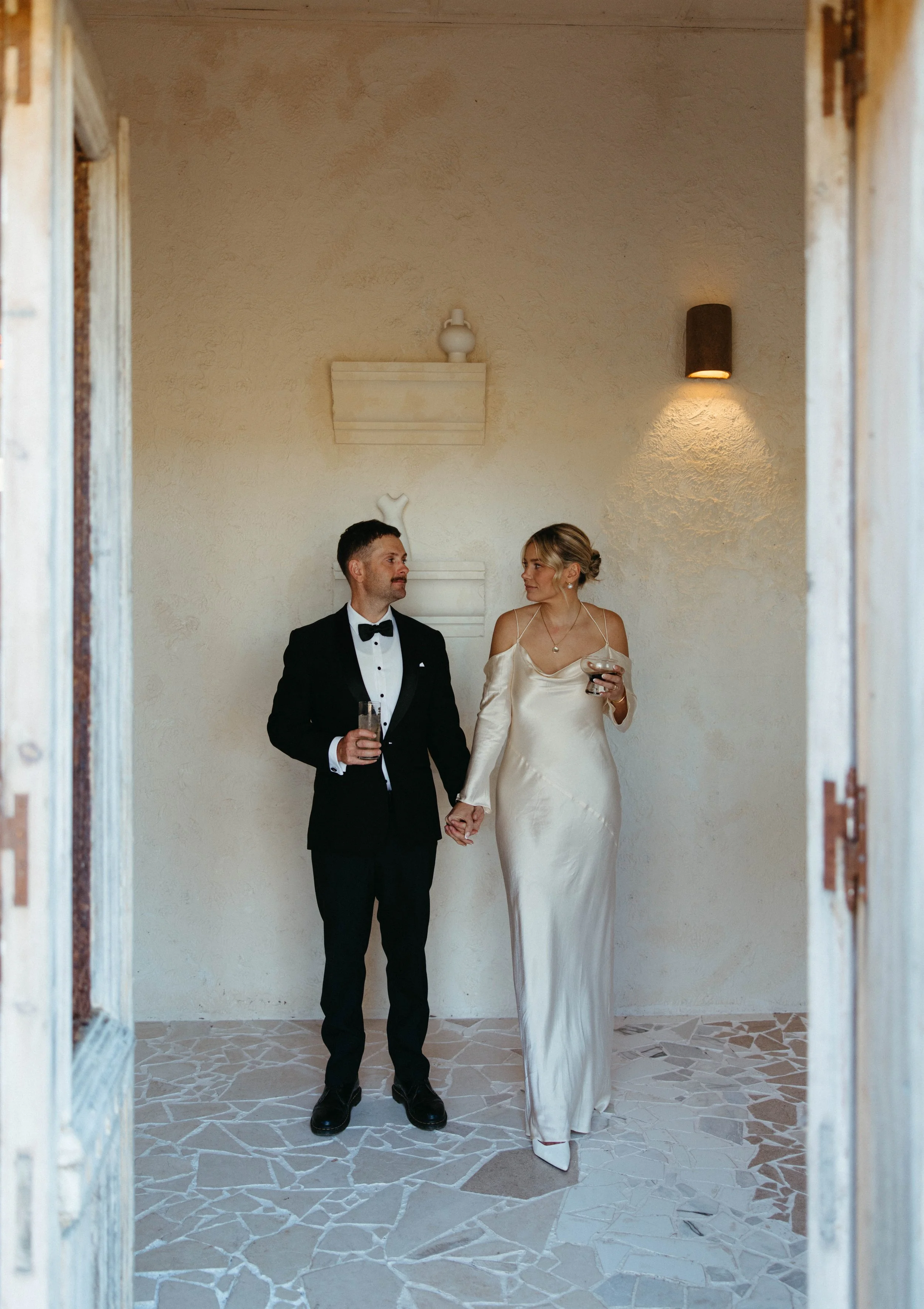 A groom and bride holding hands in an indoor setting, the groom dressed in a black tuxedo with a bow tie and the bride in a satin wedding gown, both holding drinks and looking at each other.