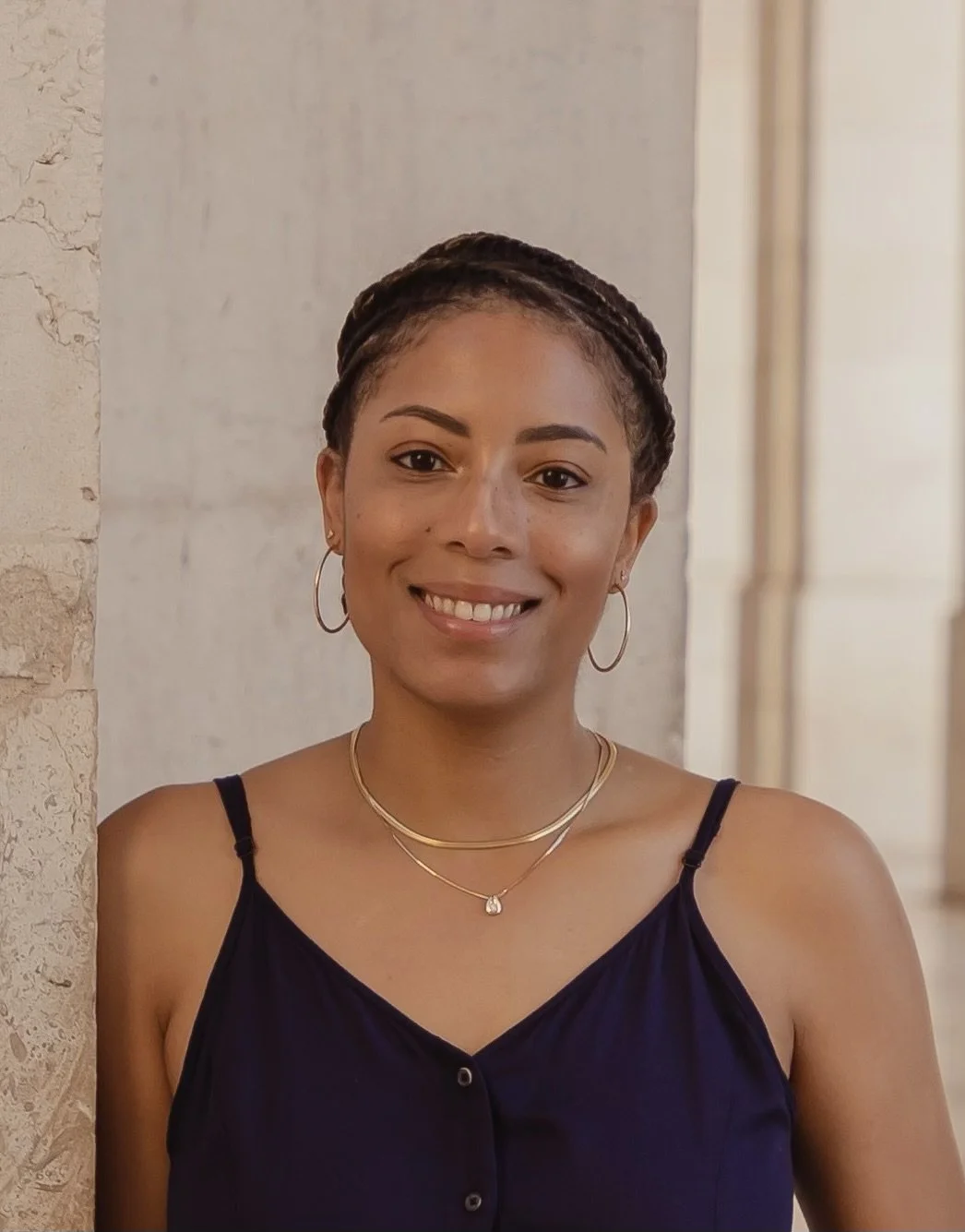 A woman smiling with short braided hair, wearing hoop earrings, layered necklaces, and a navy sleeveless top, standing against a neutral background.