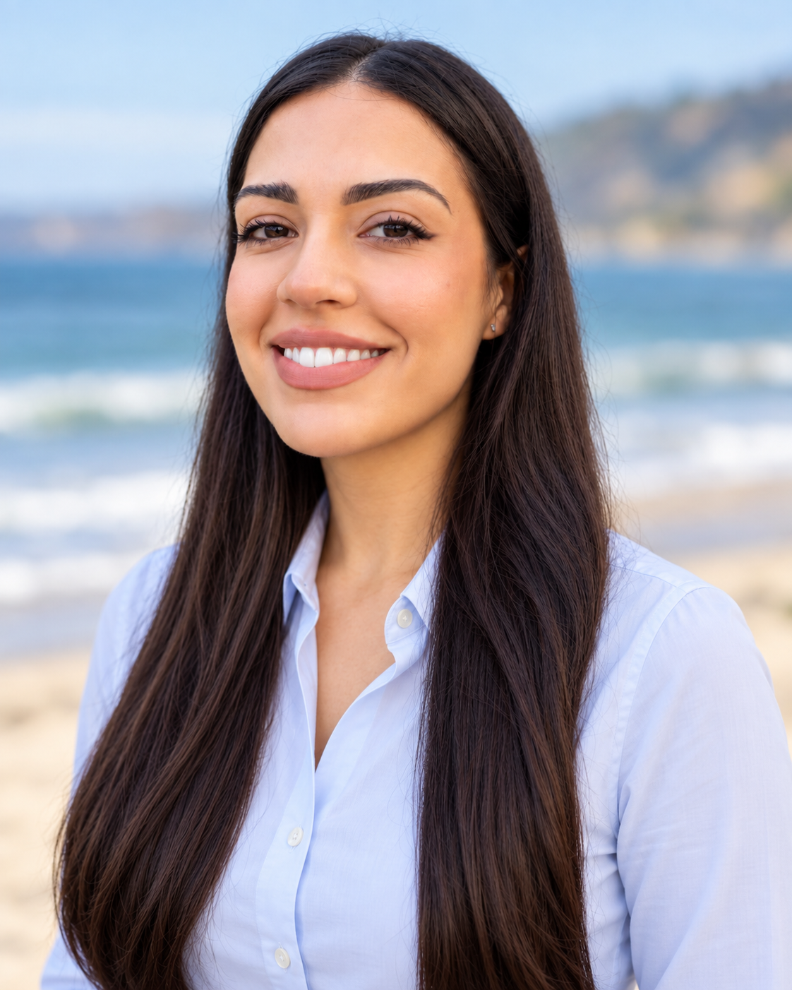 A young woman with long dark hair, wearing a light blue button-up shirt, smiling at the camera on a beach with the ocean and hills in the background.