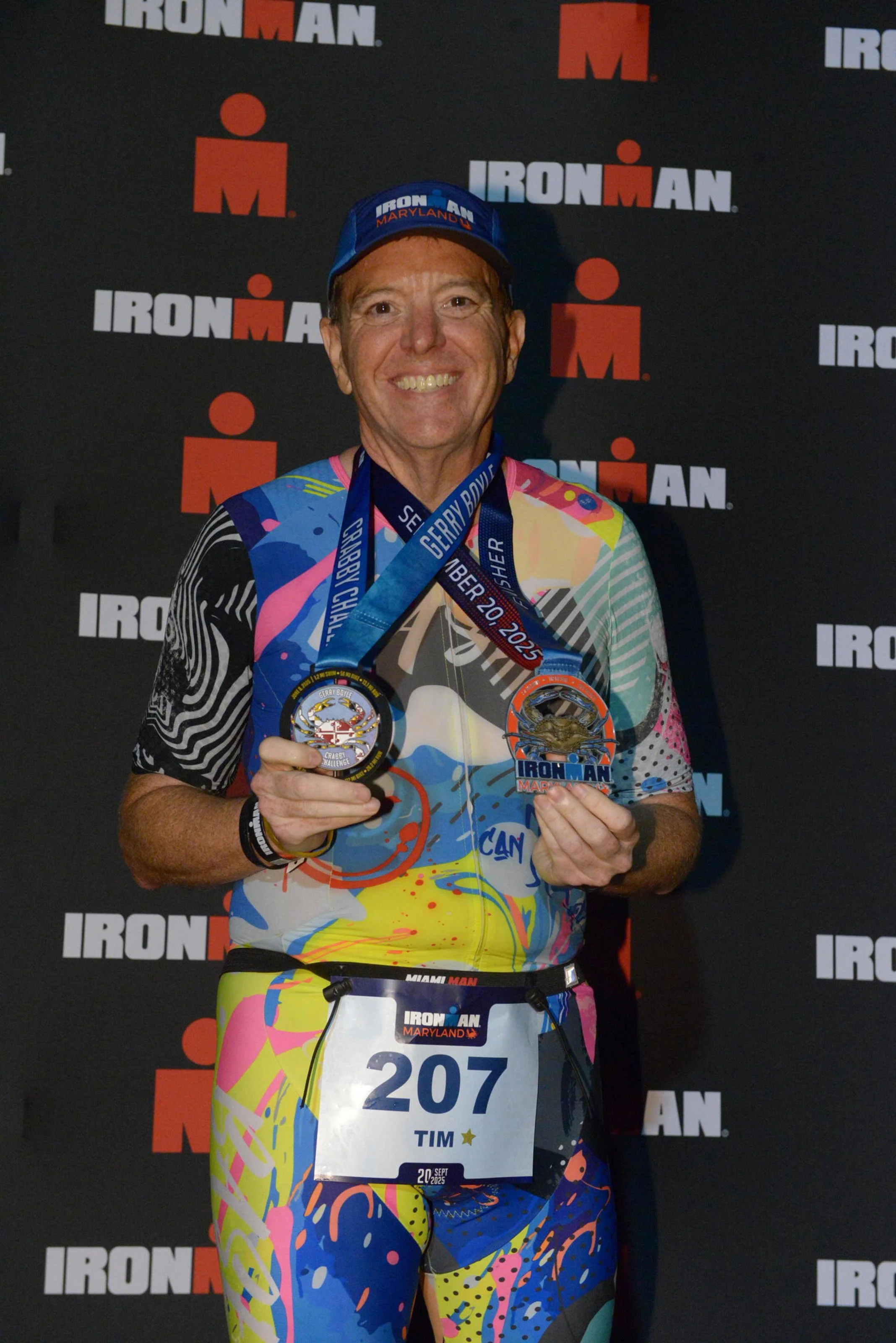 Smiling male athlete holding a finisher medal in front of a black backdrop with 'Ironman' logos, wearing a colorful athletic outfit, a blue cap, and a race bib.