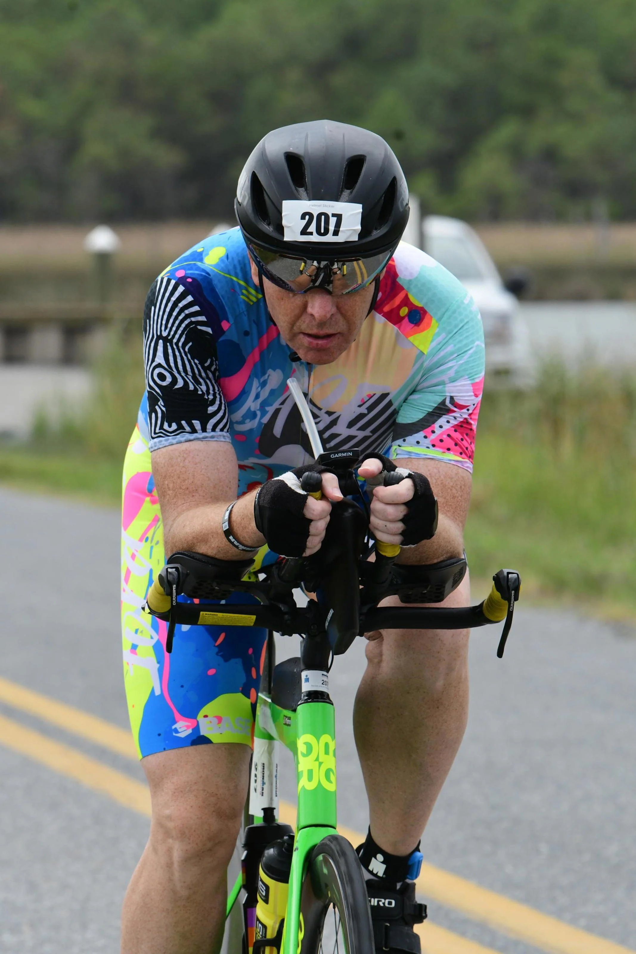 Male cyclist wearing a colorful jersey and a black helmet, riding a triathlon bike on a road.