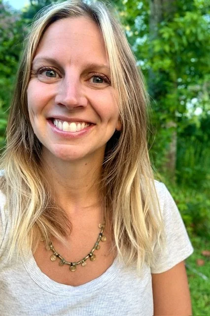 A woman with long blonde hair and blue eyes smiling outdoors with green trees in the background.