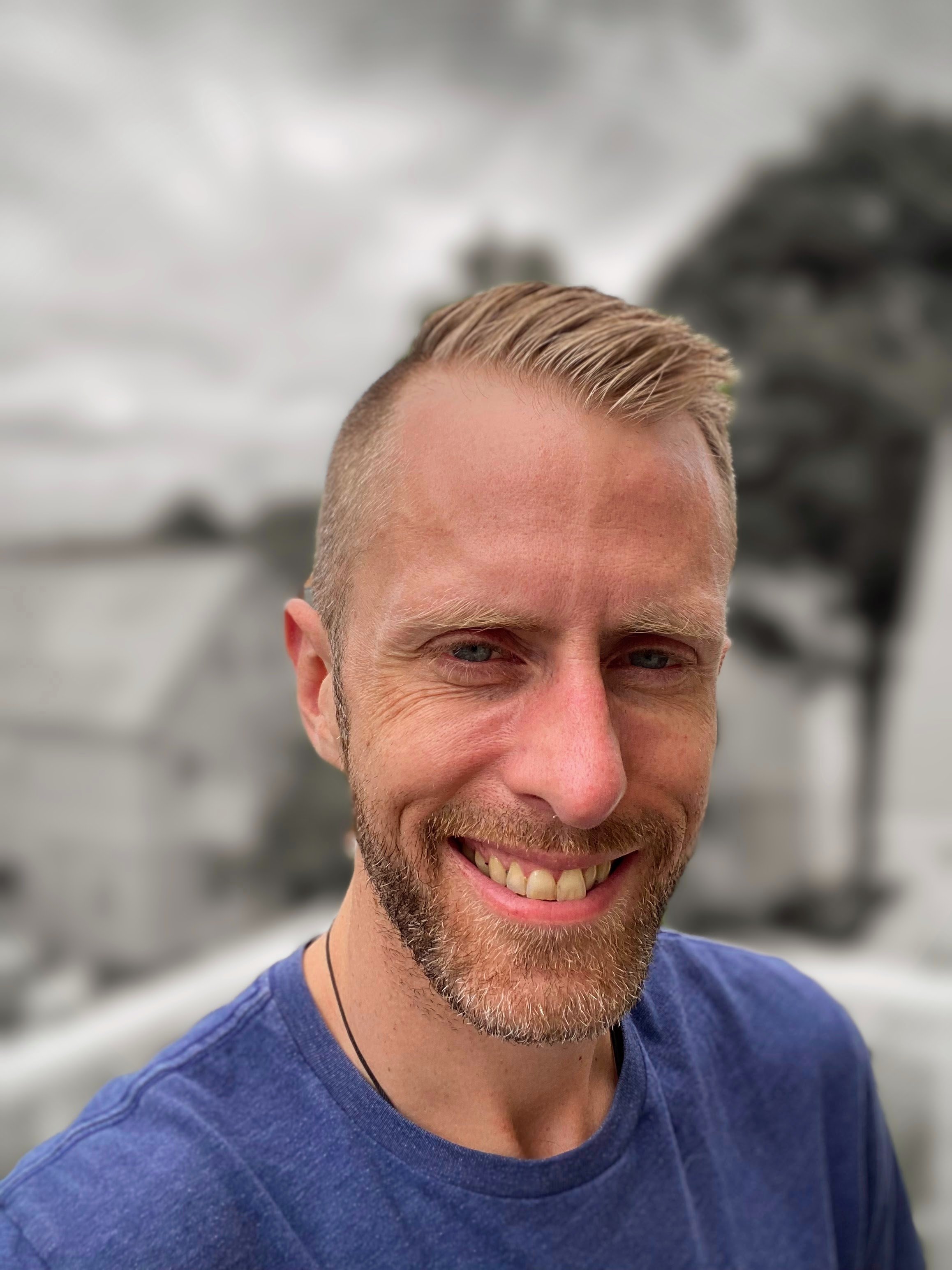 Close-up of smiling man with blonde hair, beard, wearing a blue shirt and a black necklace outdoors with blurred rocky landscape in the background.