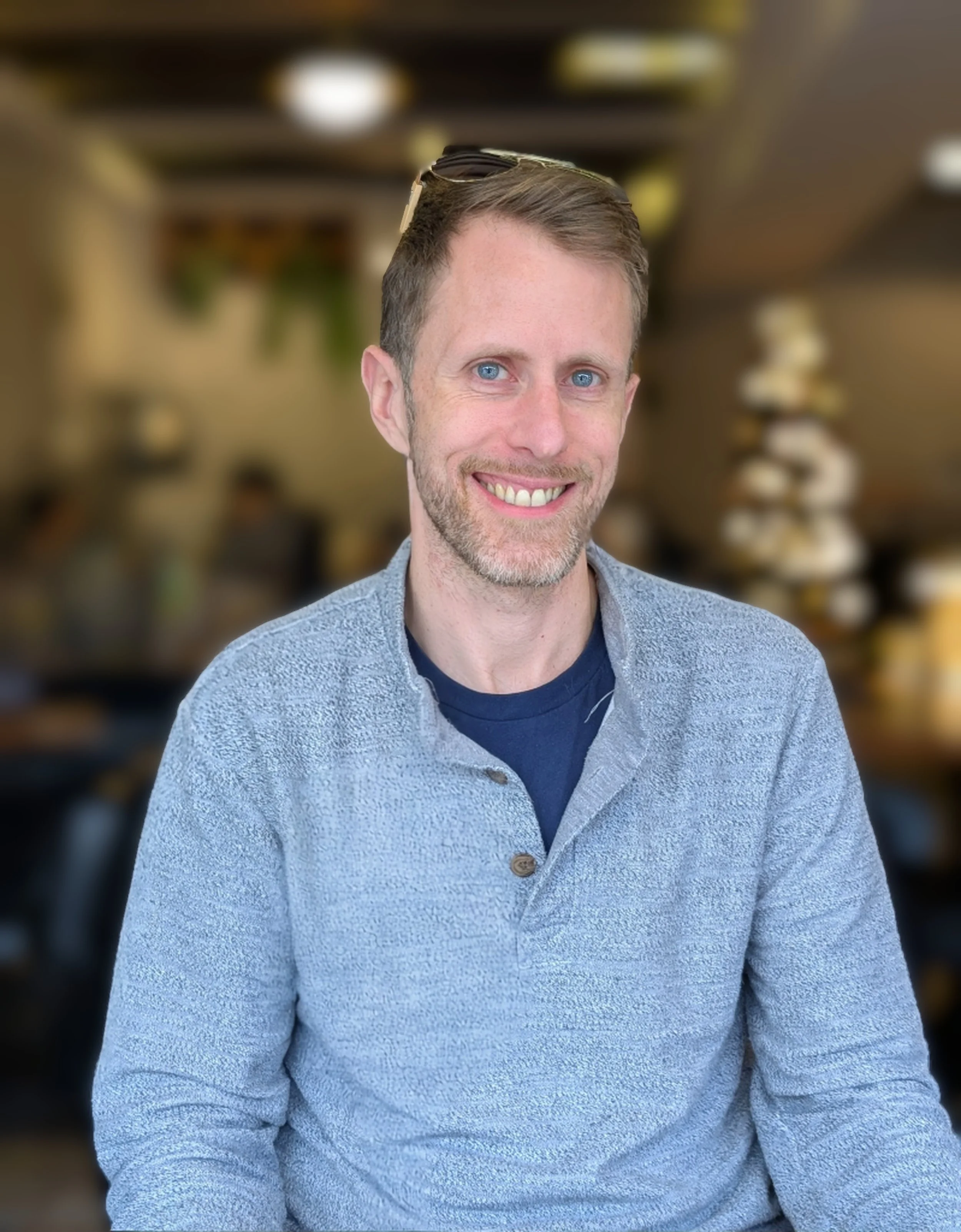 A smiling man with light brown hair and a beard, wearing a blue shirt and a gray textured sweater, in a blurred indoor setting.