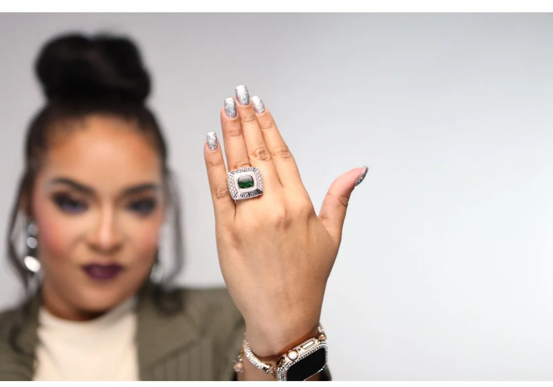 Woman displaying her hand with a large, green and silver ring, wearing glittery silver nail polish, earrings, bracelet, and smartwatch, with blurred background.