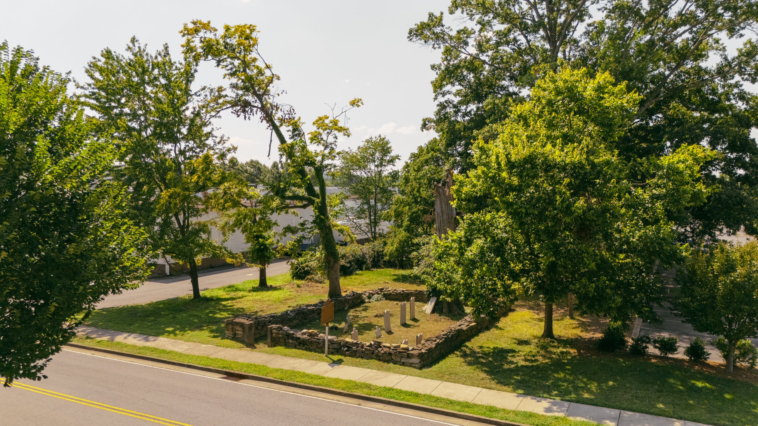 An aerial view of historic graveyard, Mallory Cemetery, enclosed by a low stone wall, located along a street with trees providing shade.