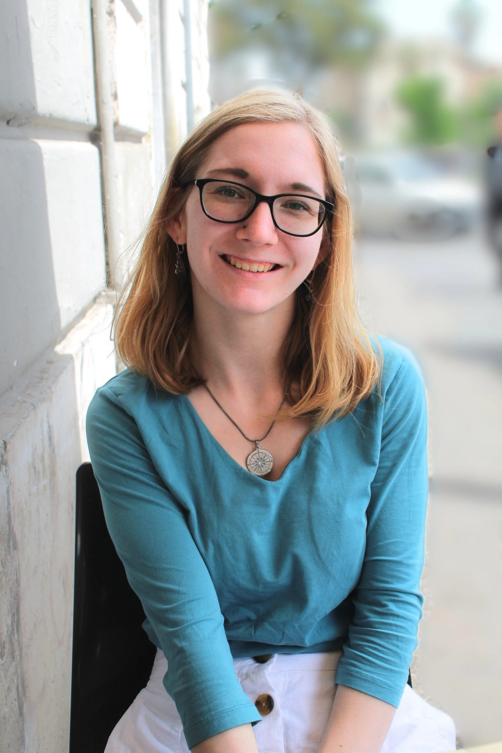 A young woman with glasses and blonde hair smiling while sitting by a window in a casual blue top with a necklace is board member Paige Hurley.