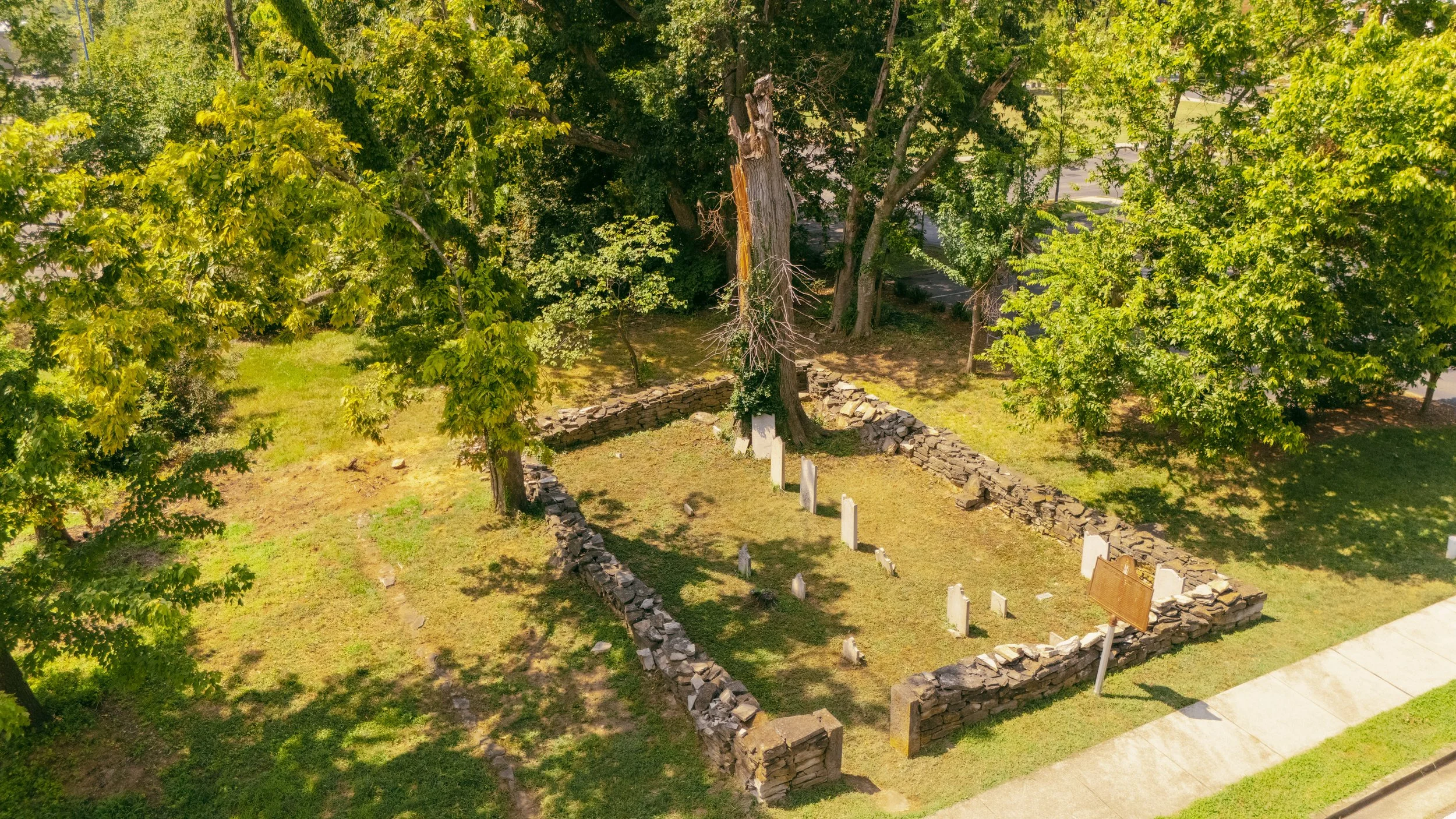 Aerial view of a small historic cemetery enclosed by stone walls, with a large tree and several headstones inside, near a sidewalk and surrounded by green trees.