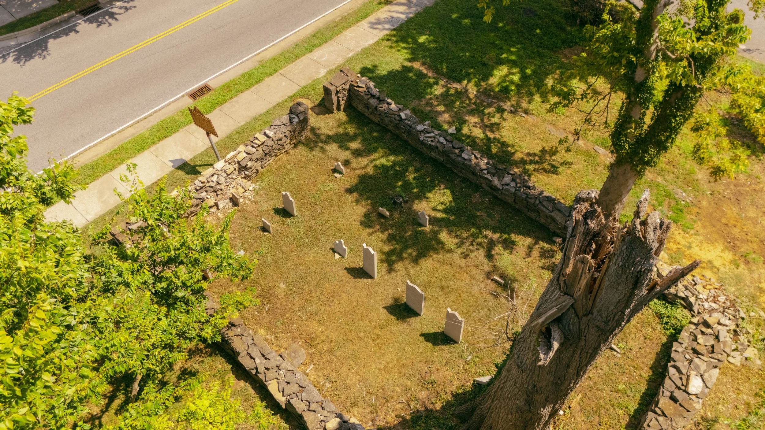 An aerial view of a small, old cemetery with stone grave markers enclosed by a stone wall, next to a sidewalk and a street with double yellow lines. There are trees casting shadows over the graves.