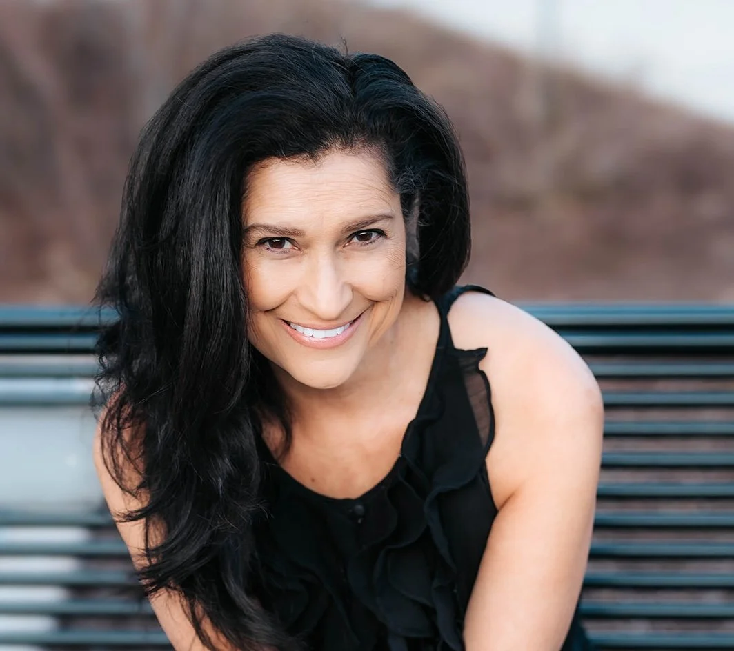 A woman with long dark hair smiling, sitting on a bench outdoors is board member Cyndi Benavides.