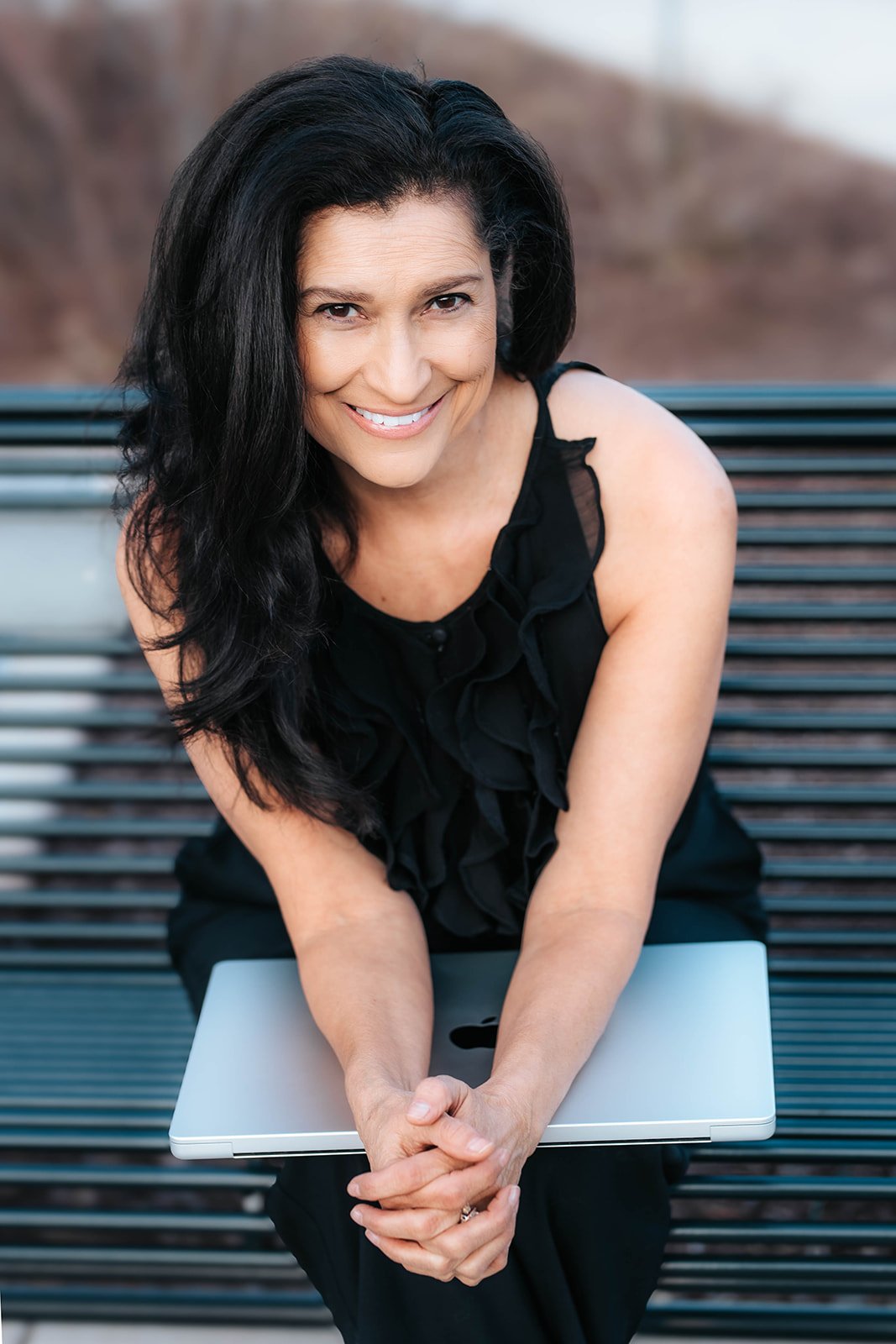 A woman with long black hair, smiling and wearing a sleeveless black top, is sitting on a park bench with her hands clasped over a closed laptop resting on her lap. The background is blurred.