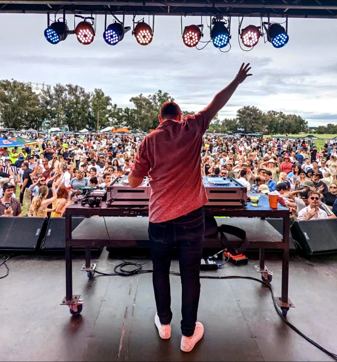 DJ on stage performing at outdoor music festival with large crowd gathered in front of stage, under cloudy sky.