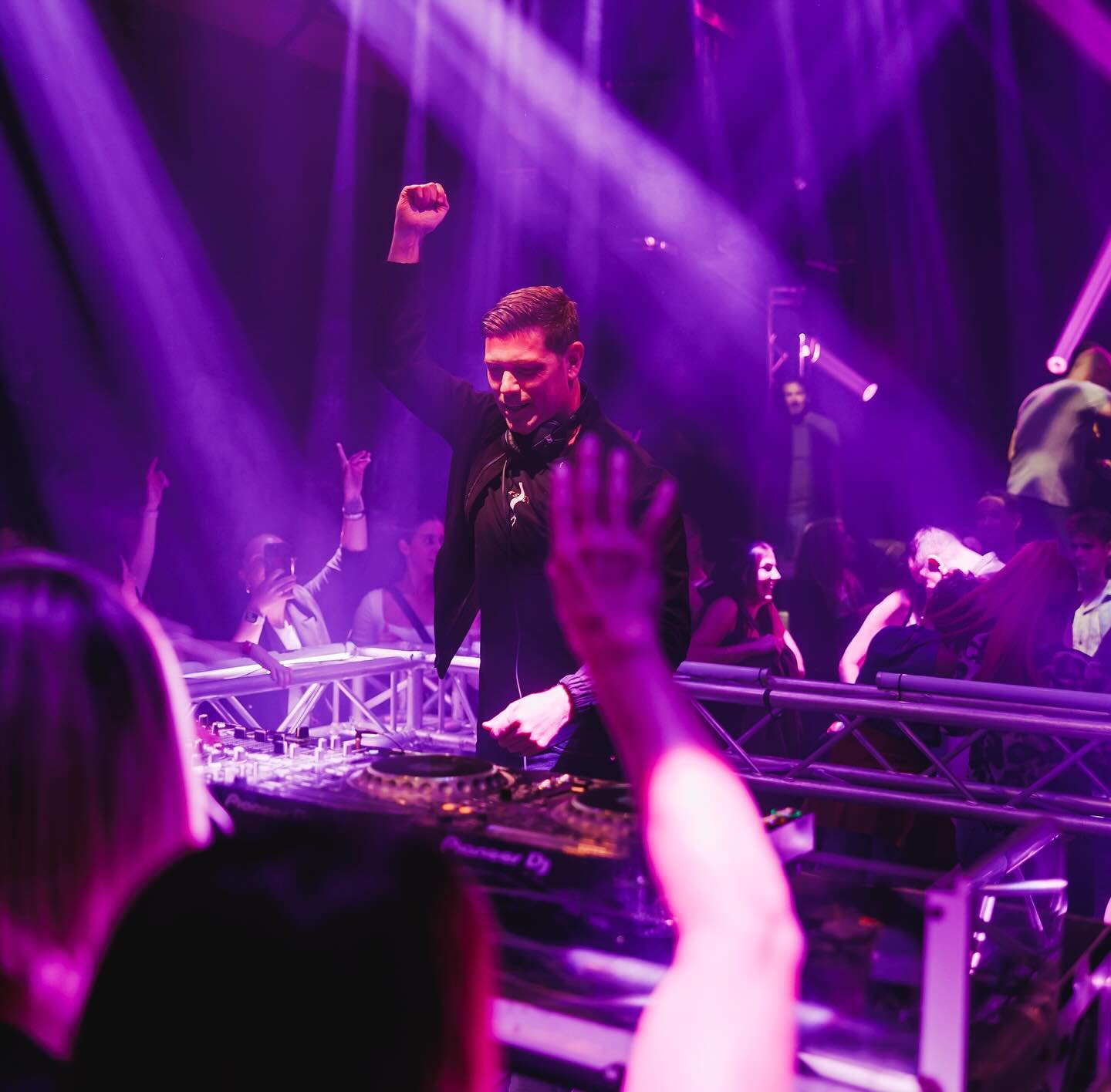 A DJ performs at a nightclub with purple lighting and an enthusiastic crowd dancing, some with hands in the air.
