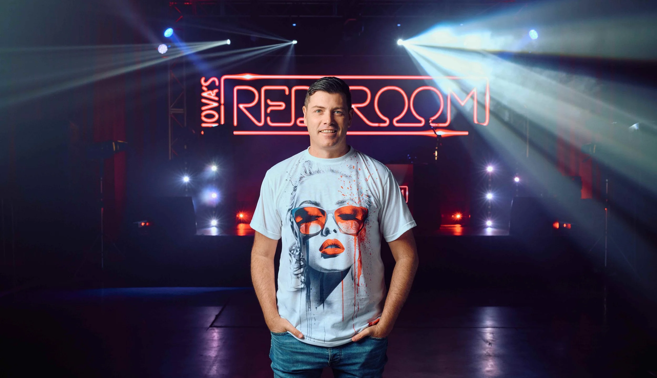 Man standing in front of a stage with neon lights and signs that read "Your Replay Room."