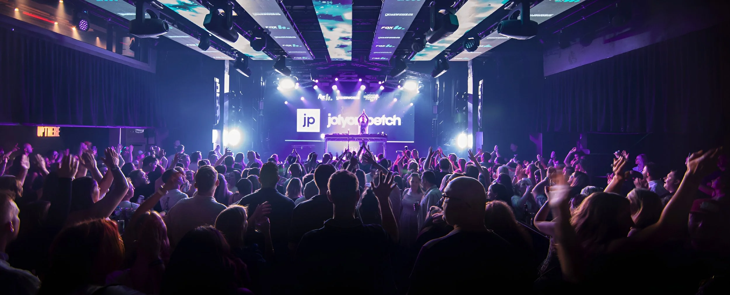 Crowd of people dancing at a concert or club with DJ on stage and bright stage lights.