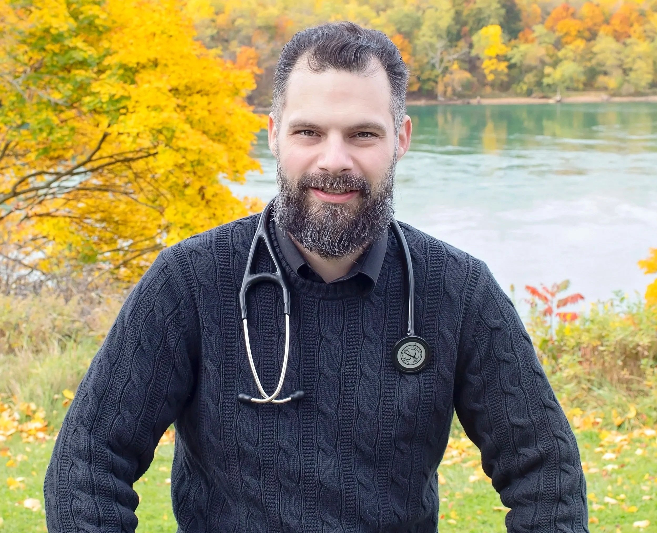 A male healthcare professional with a beard wearing a dark sweater and a stethoscope around his neck, standing outdoors during autumn by a river with yellow and orange trees in the background.