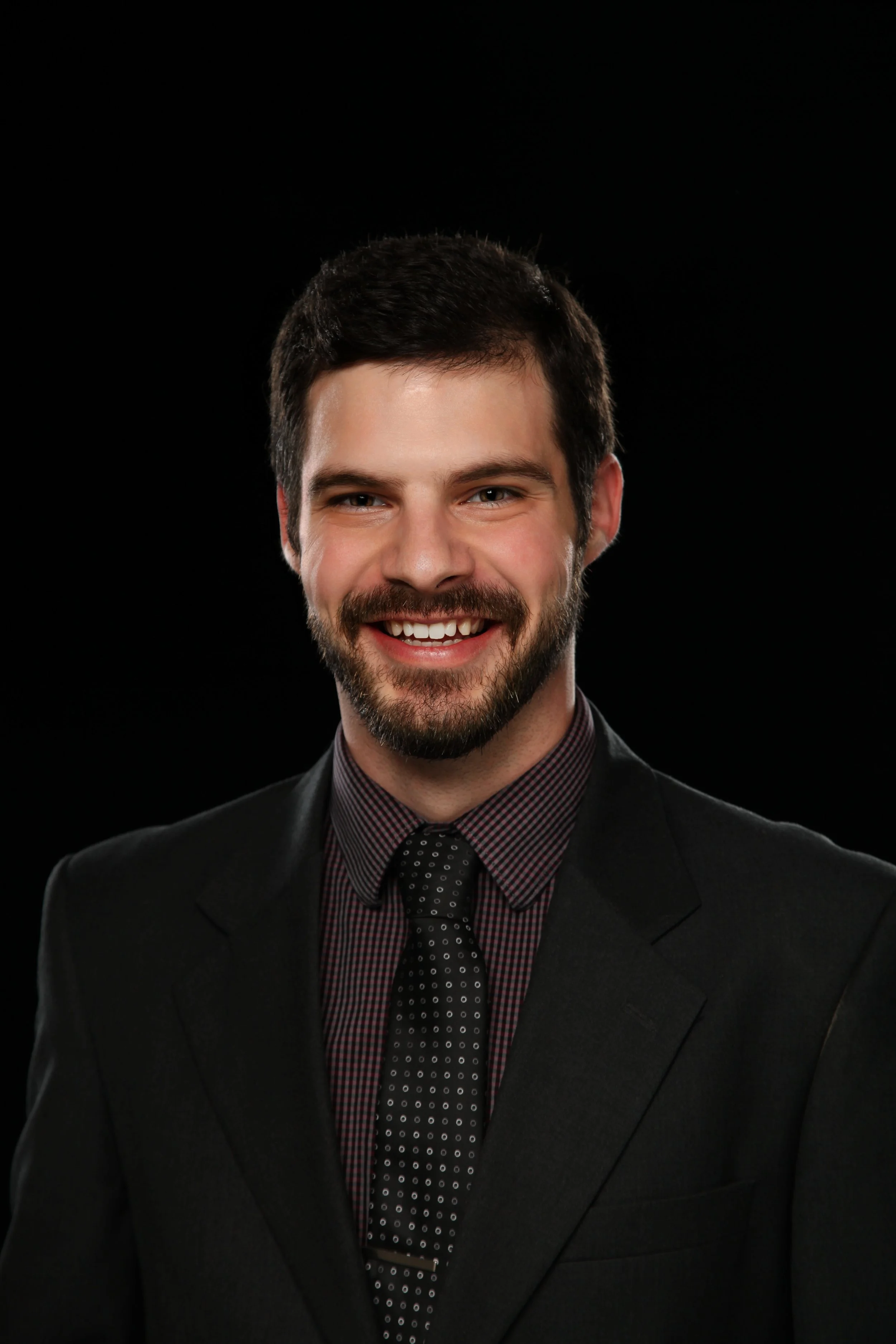 A young man with dark hair, a beard, and a mustache, dressed in a suit, shirt, and tie, smiling against a black background.