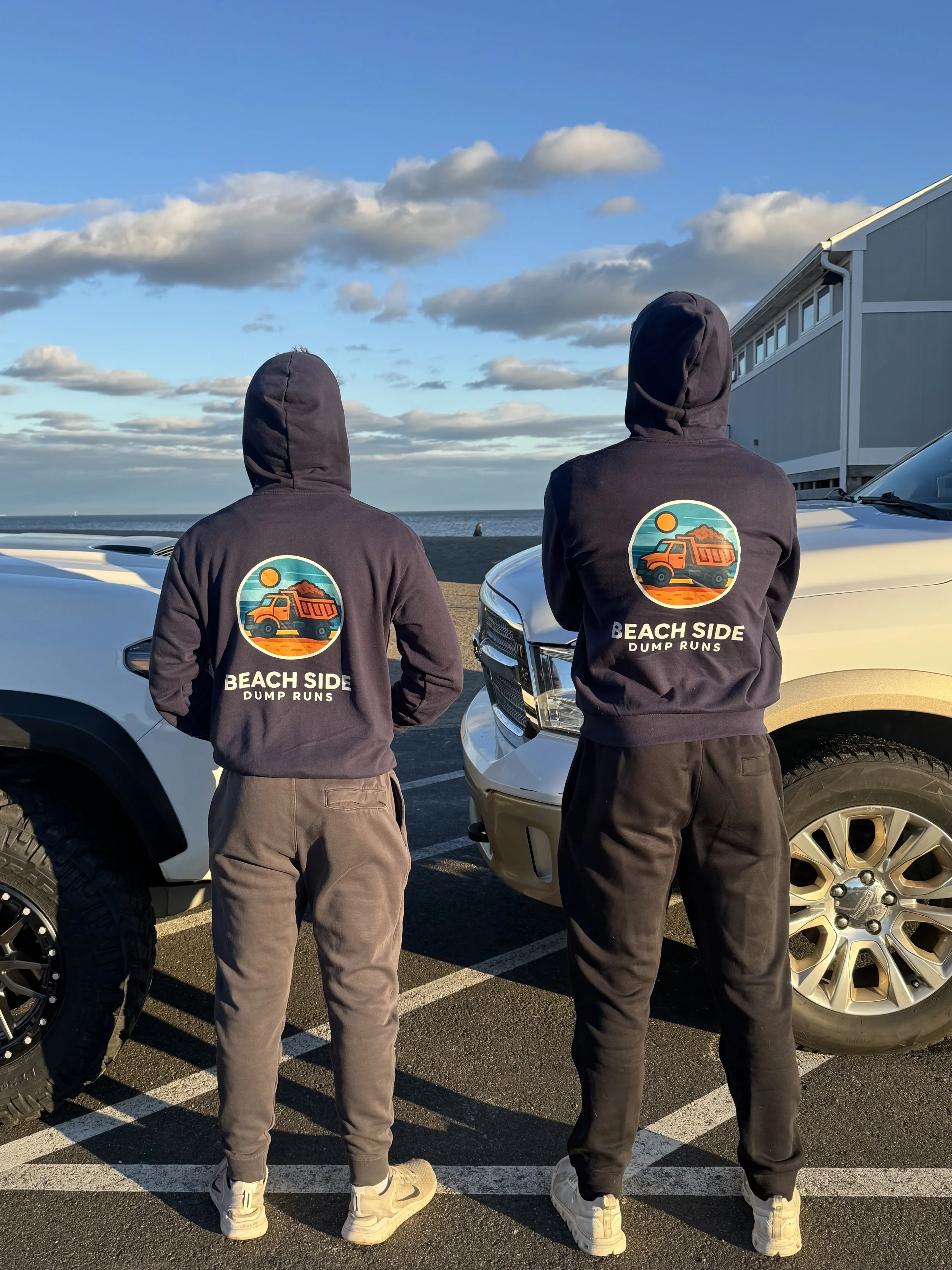 Two people standing in front of beachside parking lot wearing hoodies with a logo of a dump truck and the words 'Beach Side Dump Runs' on the back, with the ocean and sky in the background.