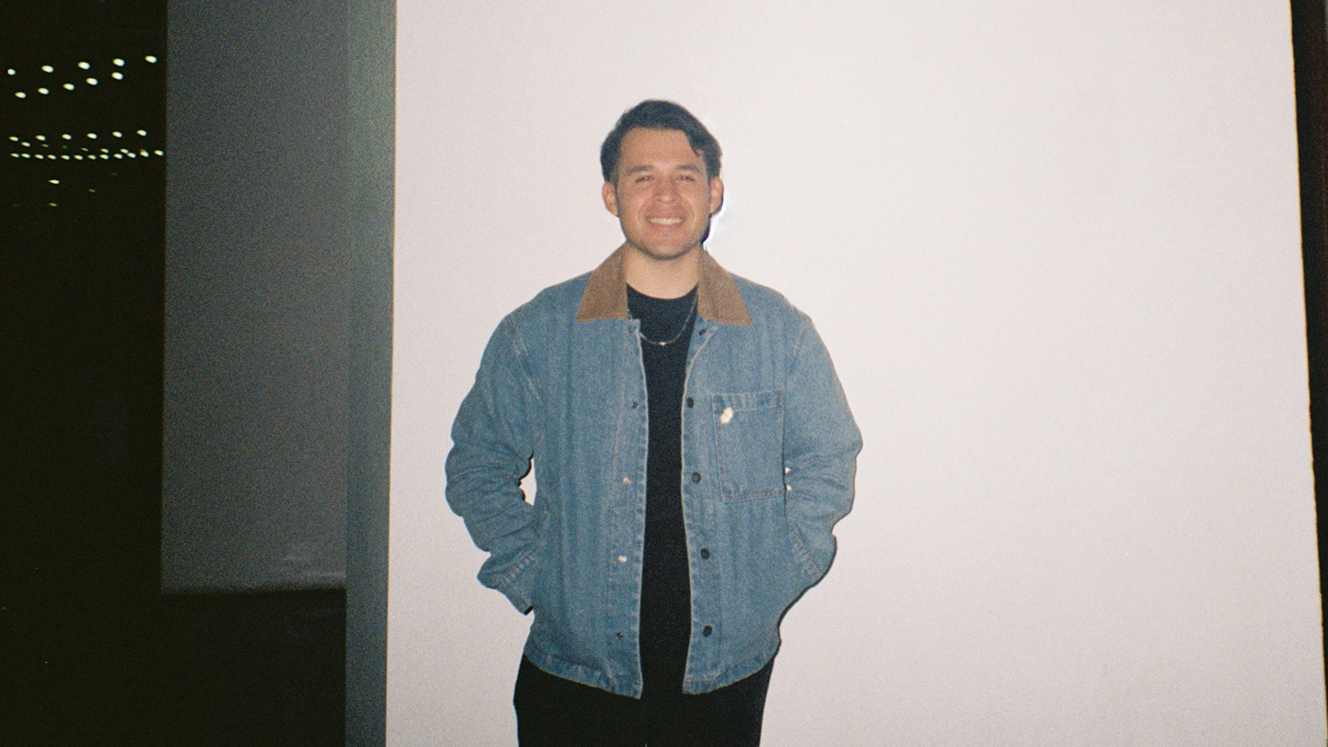 Young man standing and smiling in front of a white background, wearing a denim jacket with a brown collar and a black shirt.