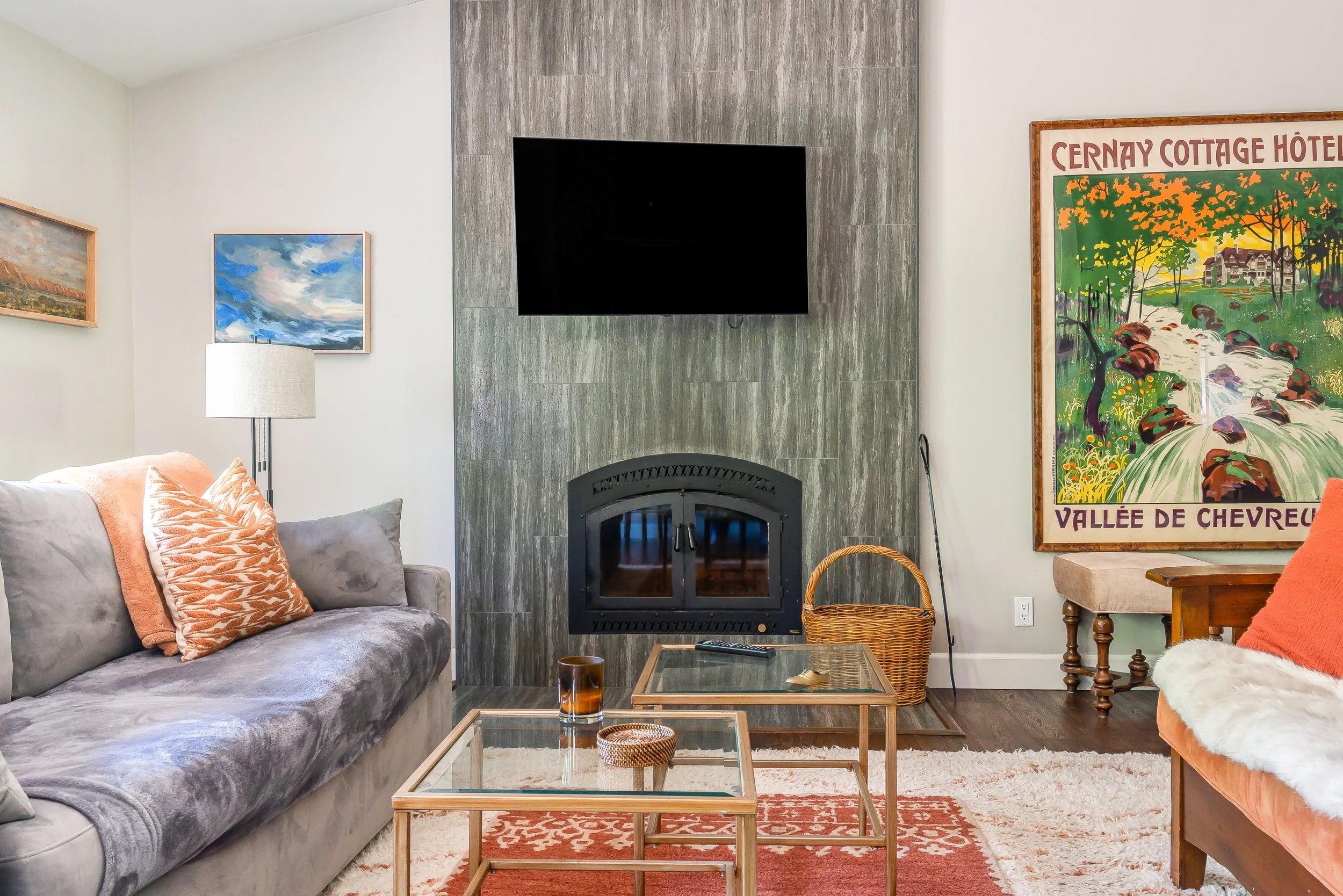 Living room with gray sofa, colorful pillows, a lamp, a wall-mounted TV above a fireplace, a glass coffee table, a woven basket, a large colorful poster on the wall, and a cream-colored armchair with an orange pillow.
