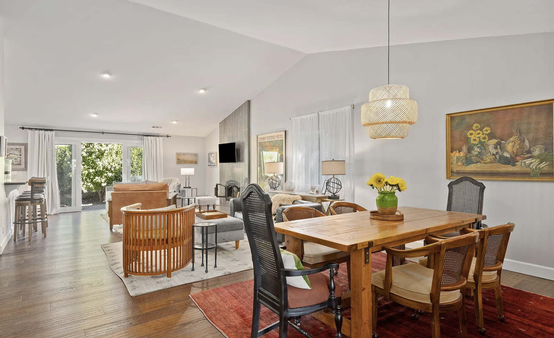 Open living and dining room with hardwood floors, white walls, natural light, sliding glass doors, and decorated with paintings, lamps, chairs, and a dining table with a sunflower centerpiece.