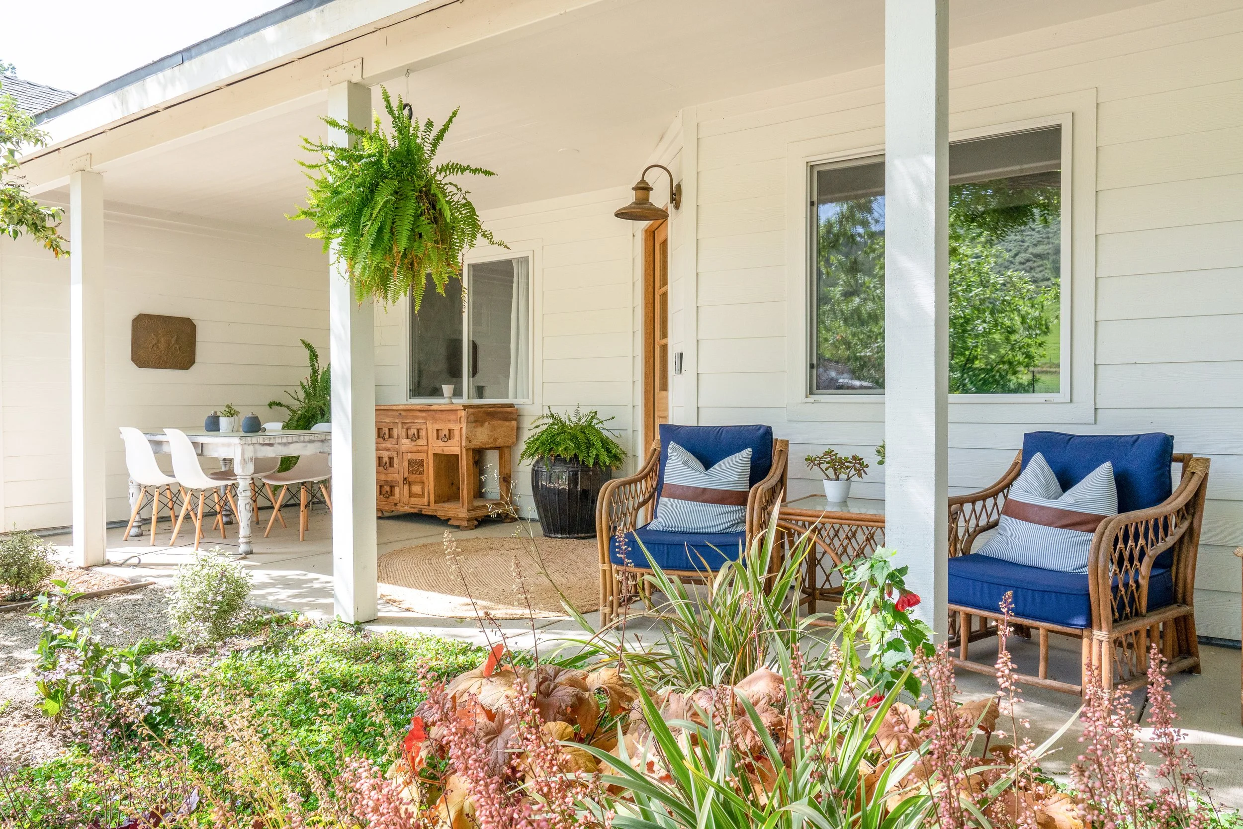 Covered porch with two wicker chairs with blue cushions and striped pillows, a small side table with a plant, a wooden bench, a dining table with white chairs, hanging plants, potted plants, and decorative items, front yard with plants and flowers.