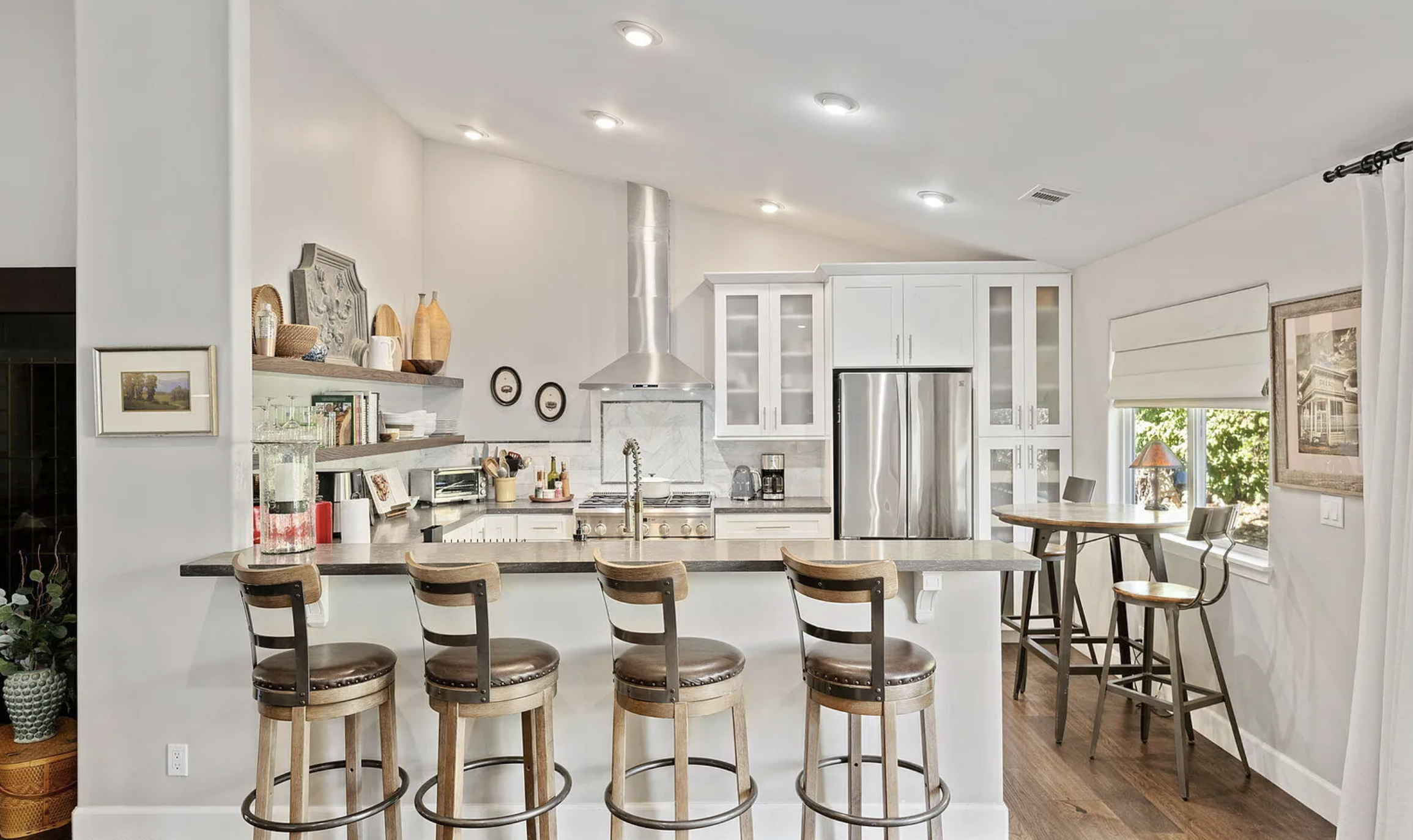 Modern kitchen with white cabinets, stainless steel appliances, and a breakfast bar with four wooden chairs. There's a small round table by the window with two chairs, and decor items on the shelves and counter. Hardwood flooring is visible.