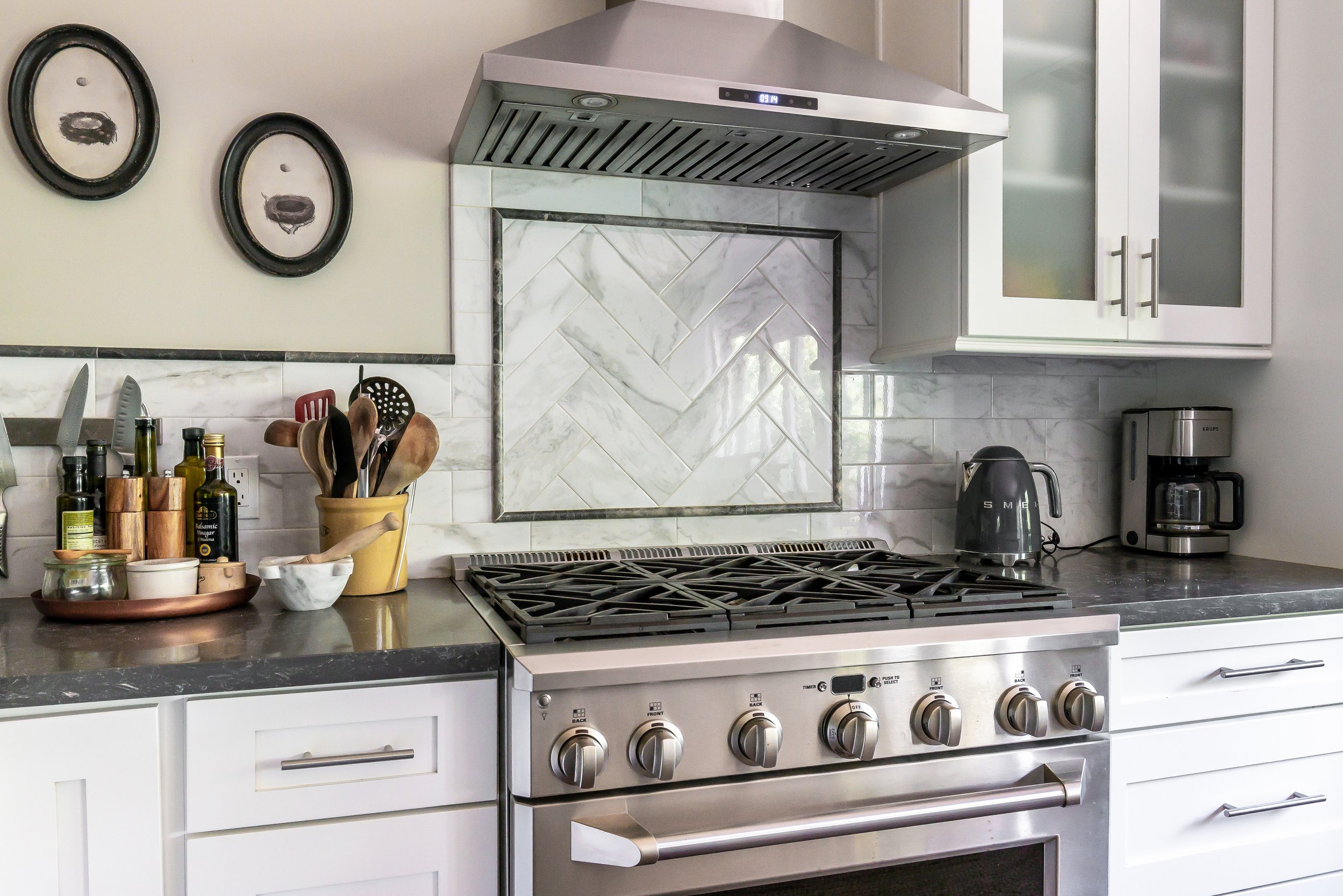 Modern kitchen with stainless steel stove, white cabinets, black countertops, and a marble backsplash. Kitchen tools and ingredients are on the counter, and two decorative wall clocks are on the wall.