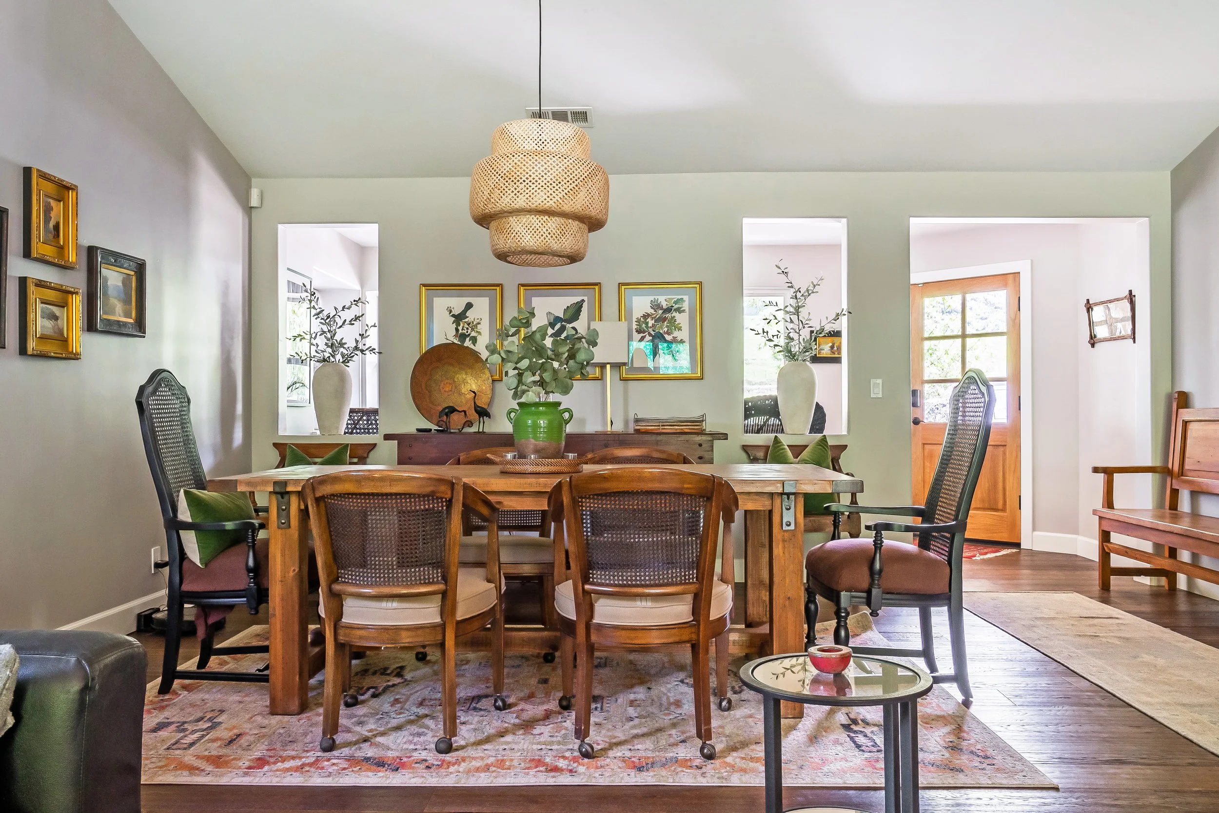 A cozy dining room with a wooden table surrounded by vintage-style chairs, a pendant light hanging above, and decorative plants and artwork on the walls.
