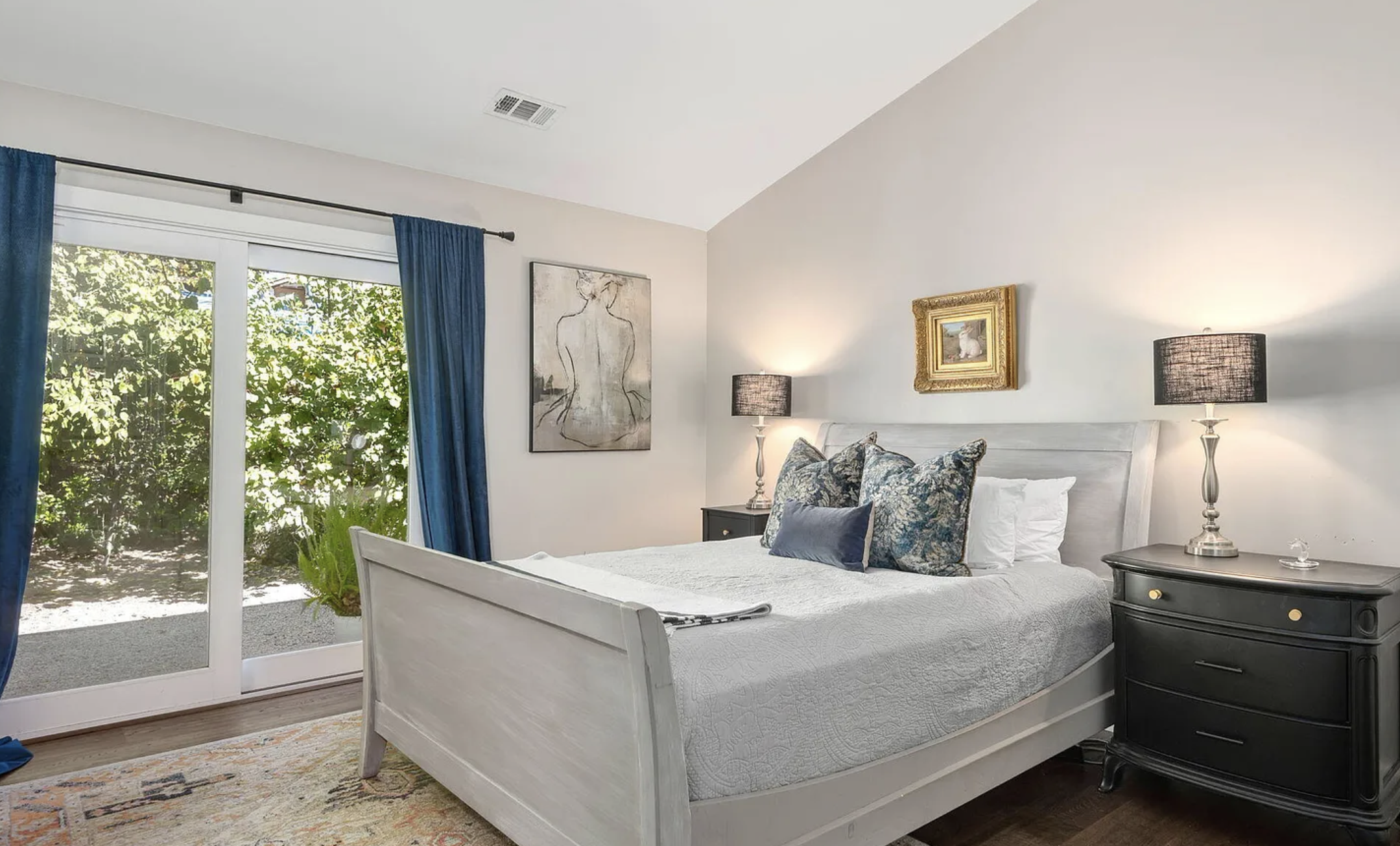 A bedroom with a white bed and decorative pillows, two black nightstands with lamps, a sliding glass door with blue curtains, and wall art, with green foliage outside.