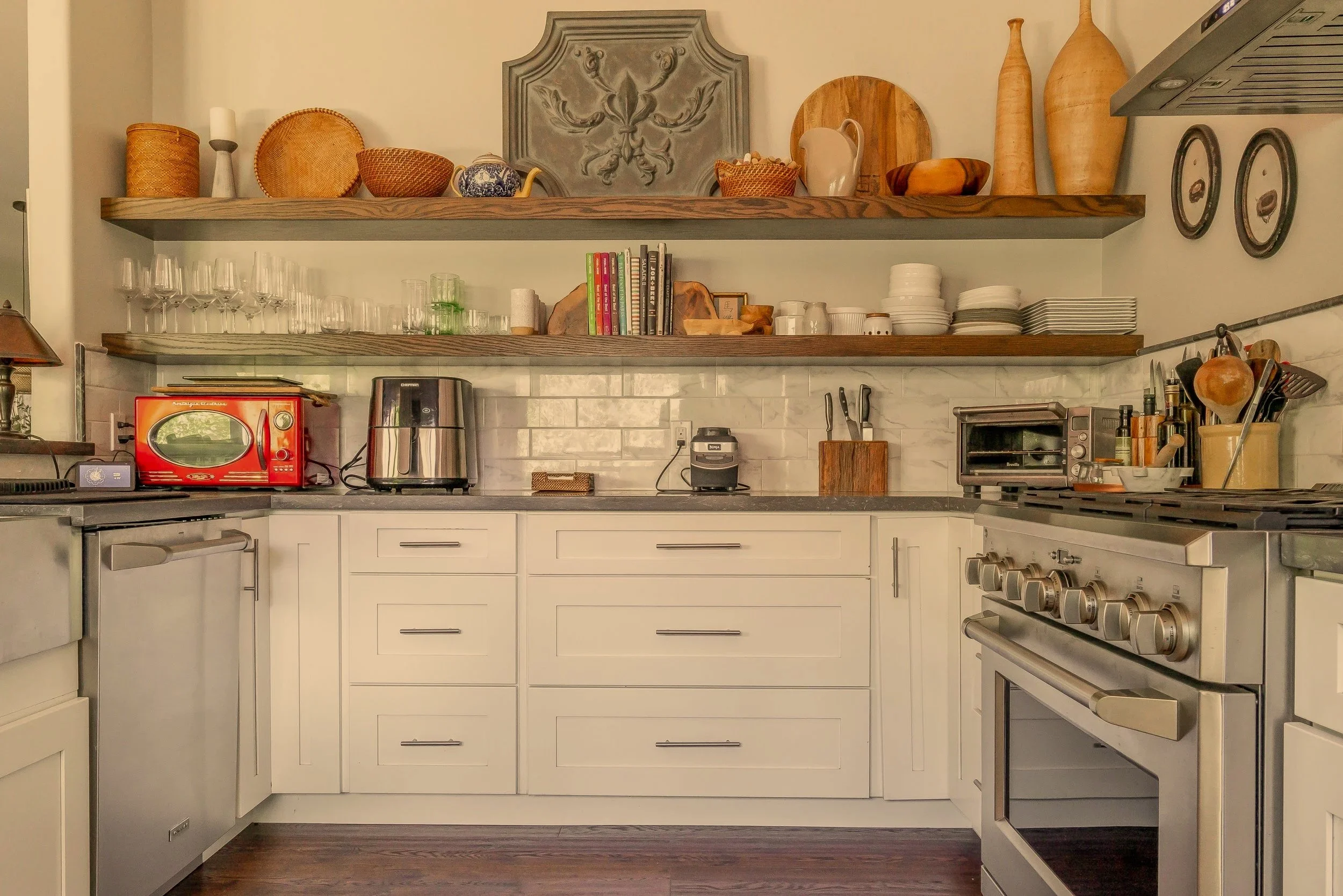 Kitchen with white cabinets, gray countertops, open wooden shelves with decorative bowls, glasses, books, and plates, stainless steel appliances, and wooden utensils.