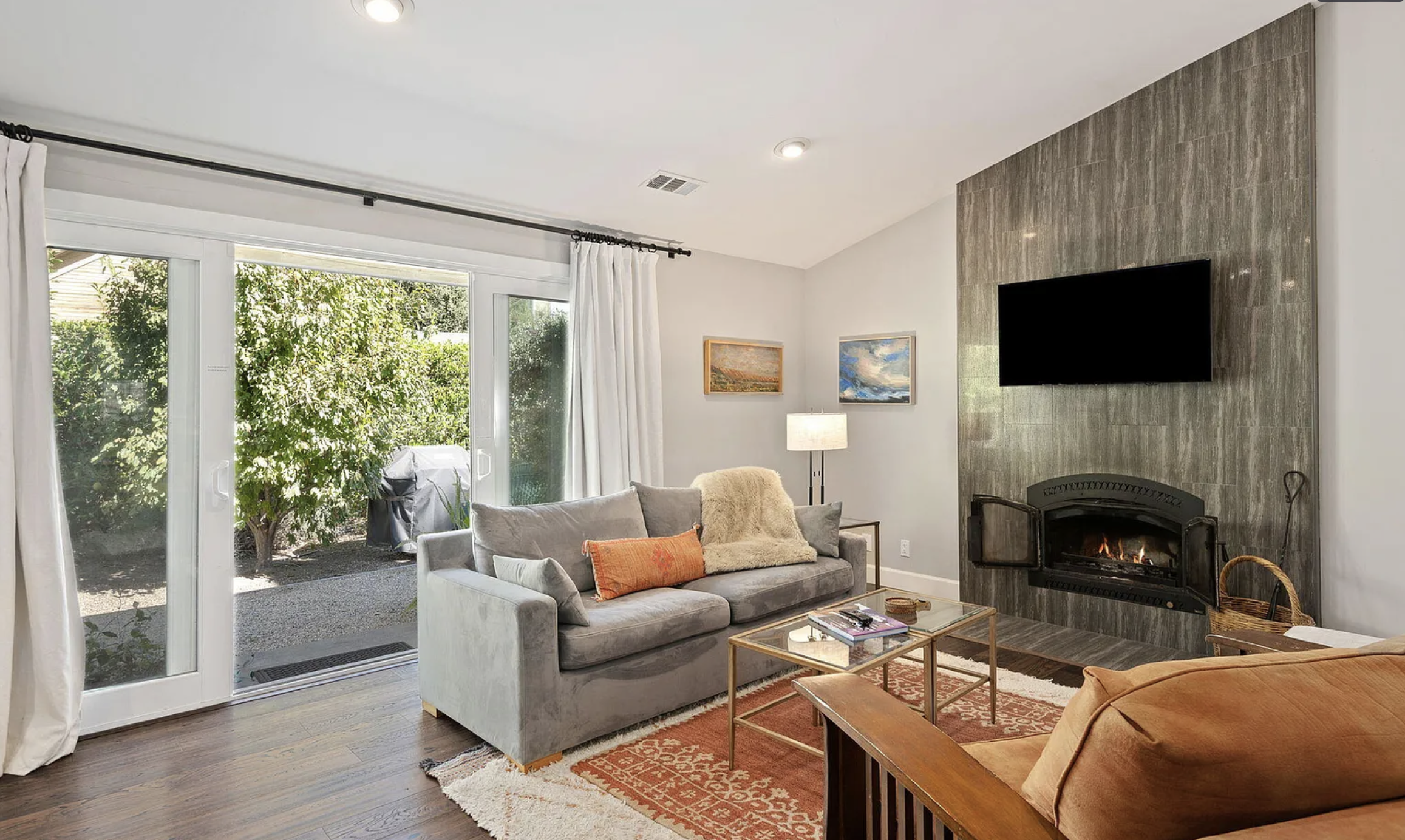 Living room with sliding glass doors, a gray sofa with orange and beige pillows, a coffee table, an orange armchair, a fireplace with a mounted black TV, and outside greenery visible through the doors.